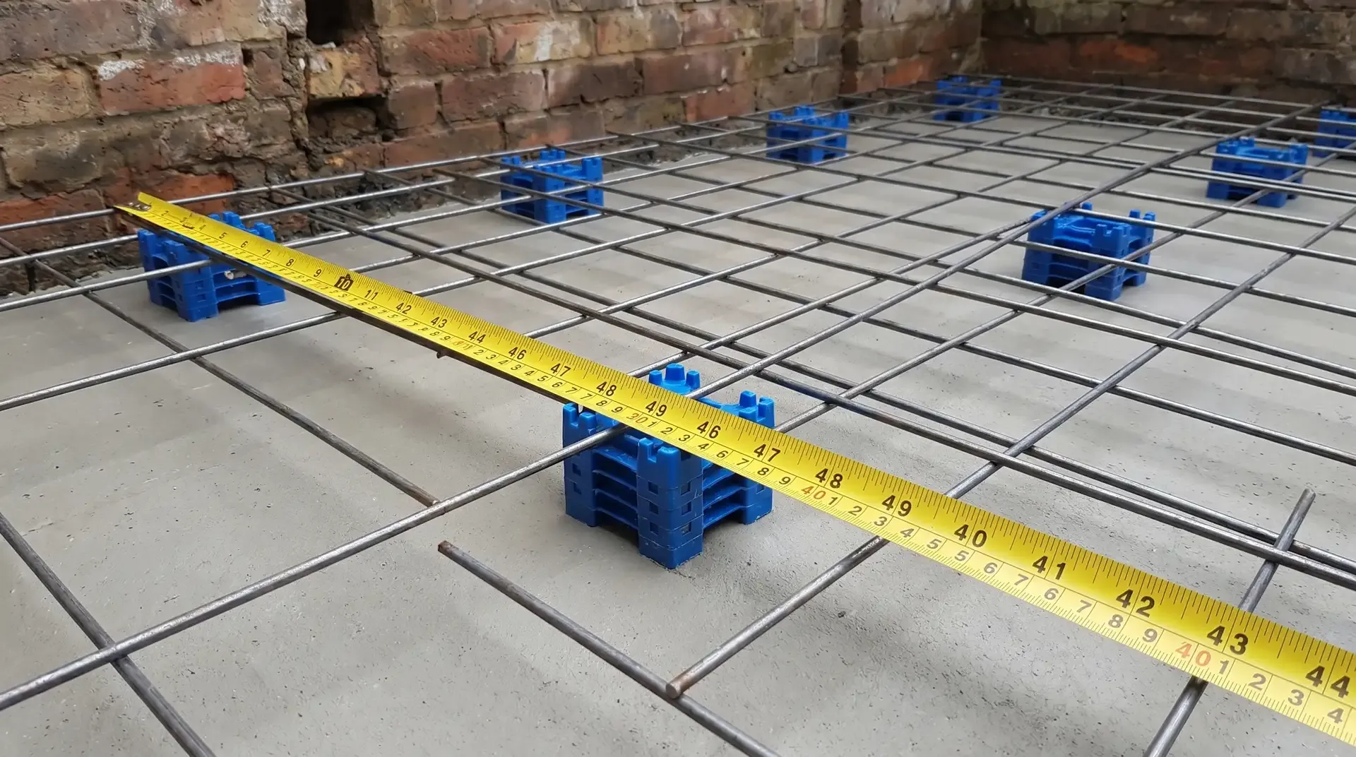 Close-up of steel reinforcement mesh resting on plastic castle spacers on top of a cured grey blinding concrete layer inside an extension footprint, with a tape measure showing approximately 40mm of cover between the mesh and the blinding surface.
