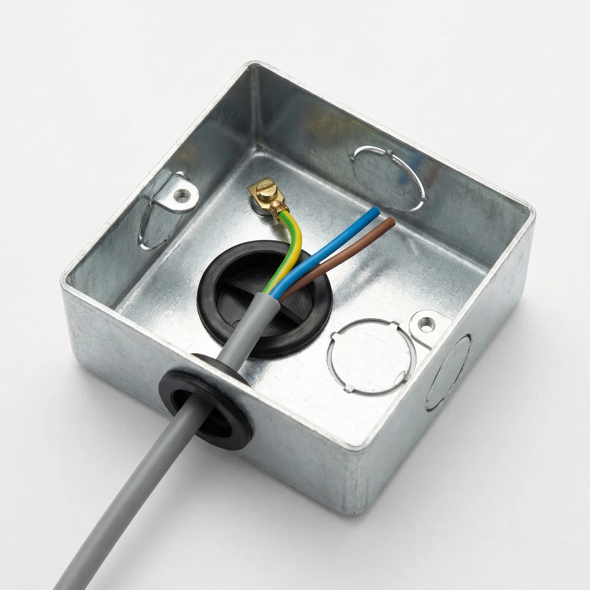 Close-up overhead photograph of a galvanised steel single gang back box with a black PVC grommet fitted in one punched-out knockout hole, a grey twin-and-earth cable passing through it, a brass earth terminal tag on the interior wall with green/yellow sleeved earth wire attached, and an adjacent un-grommeted knockout showing its sharp metal edge.