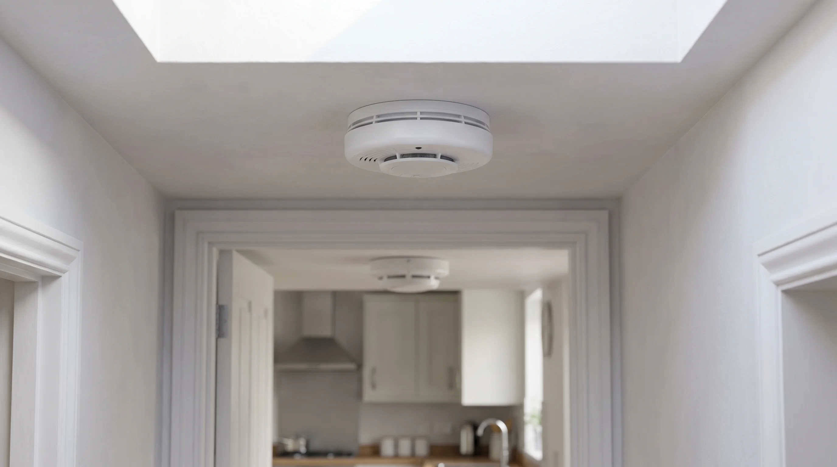 A mains-powered interlinked optical smoke alarm mounted on a hallway ceiling next to a pendant light fitting