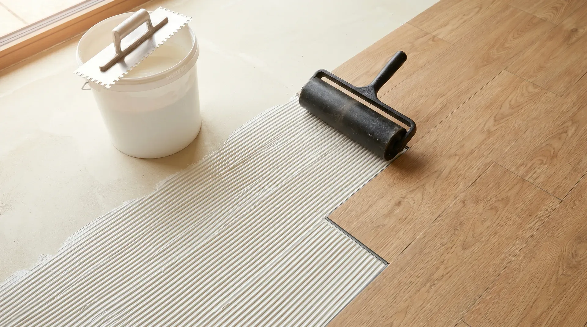 Photorealistic overhead view of an LVT glue-down installation in progress, showing fine-notched trowel spreading pressure-sensitive adhesive across a primed screed with the first row of wood-effect vinyl planks being laid