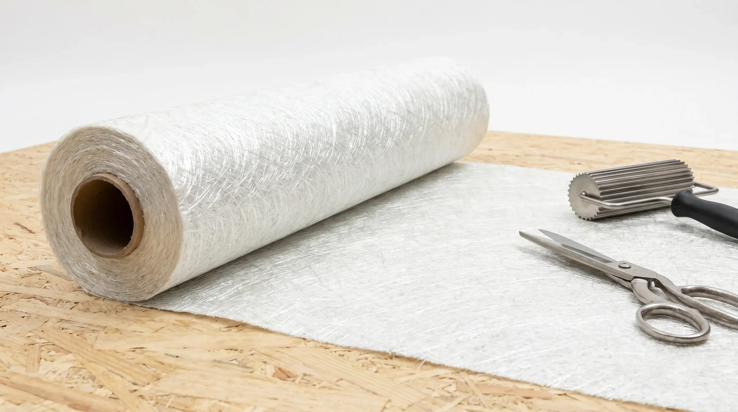 Roll of 450g chopped strand mat partially unrolled on an OSB3 flat roof deck, with the white random-fibre weave clearly visible alongside scissors and a paddle roller