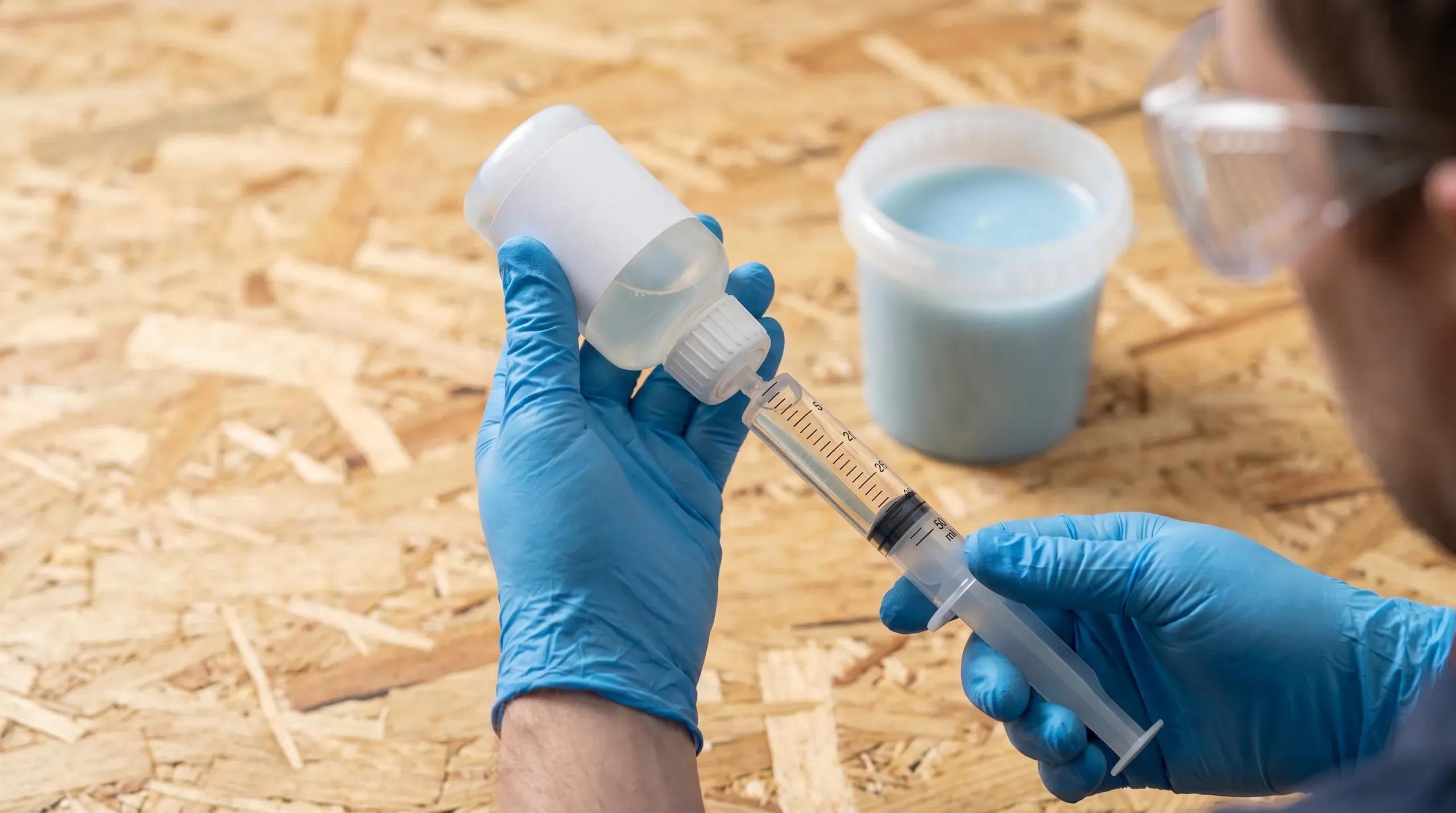 Roofer measuring MEKP catalyst from a small bottle into a bucket of polyester resin using a calibrated 50ml medical syringe, with PPE goggles and chemical-resistant gloves visible