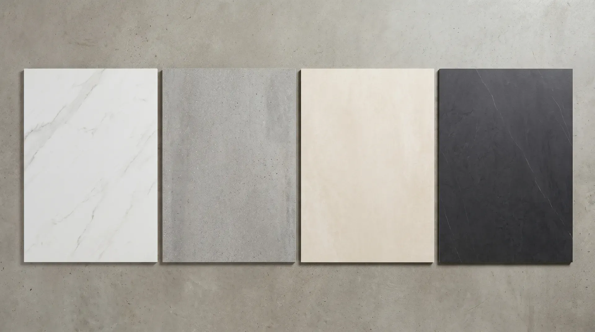 Four sintered stone slab samples laid flat on a neutral concrete studio surface, shown side by side: a white marble-effect sample, a mid-grey concrete-effect sample, a warm cream through-coloured sample, and a deep charcoal sample with subtle veining.