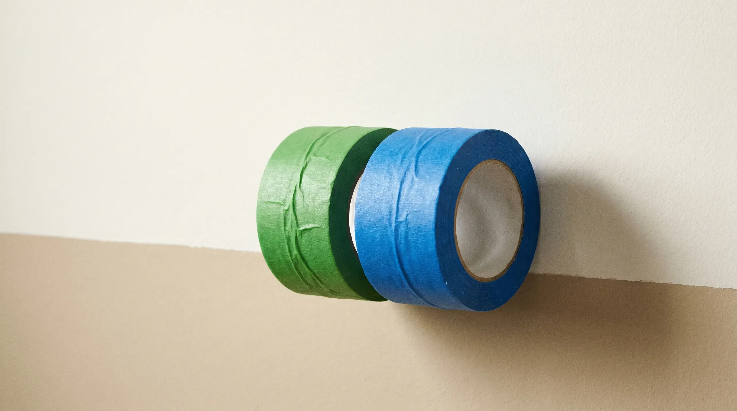A roll of green FrogTape and a roll of blue 3M Scotch Blue tape resting on a freshly painted wall with a sharp paint line visible