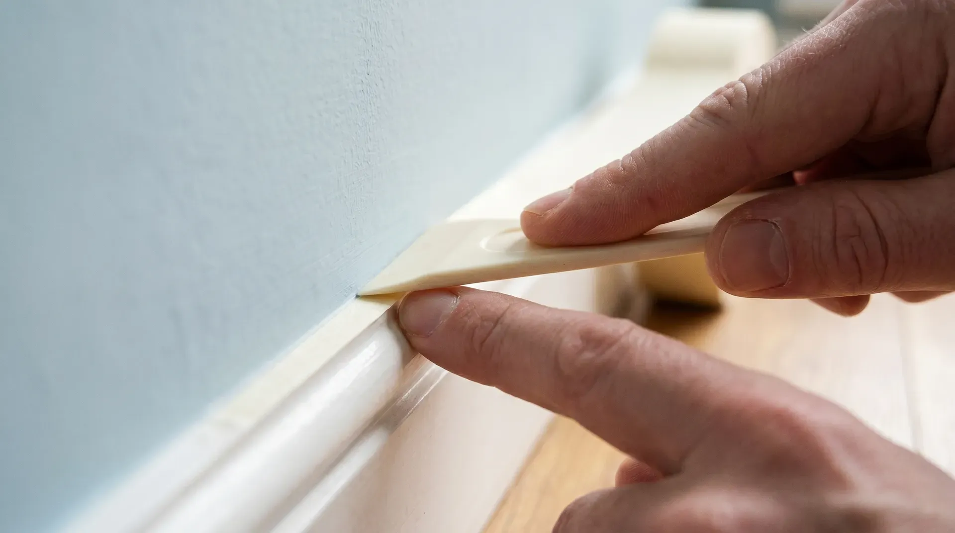 Plastic scraper pressed firmly along the edge of cream masking tape on a skirting board to seal it for a sharp paint line.