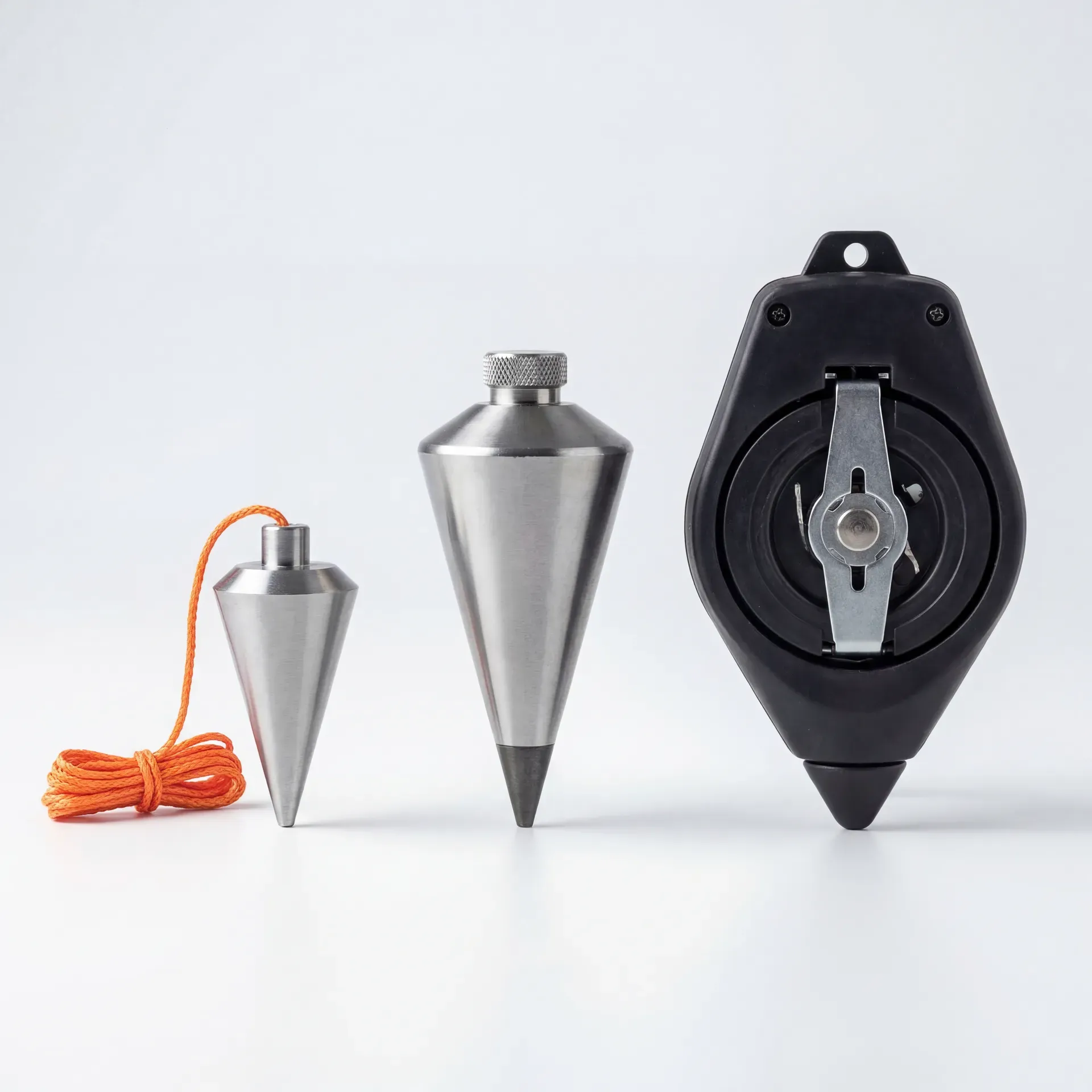 Three plumb bobs arranged left to right from smallest to largest: a small budget steel bob with orange nylon line, a mid-weight steel bob with knurled cap, and a Tajima Plumb-Rite self-stabilising unit in its black elastomer case with visible line reel.