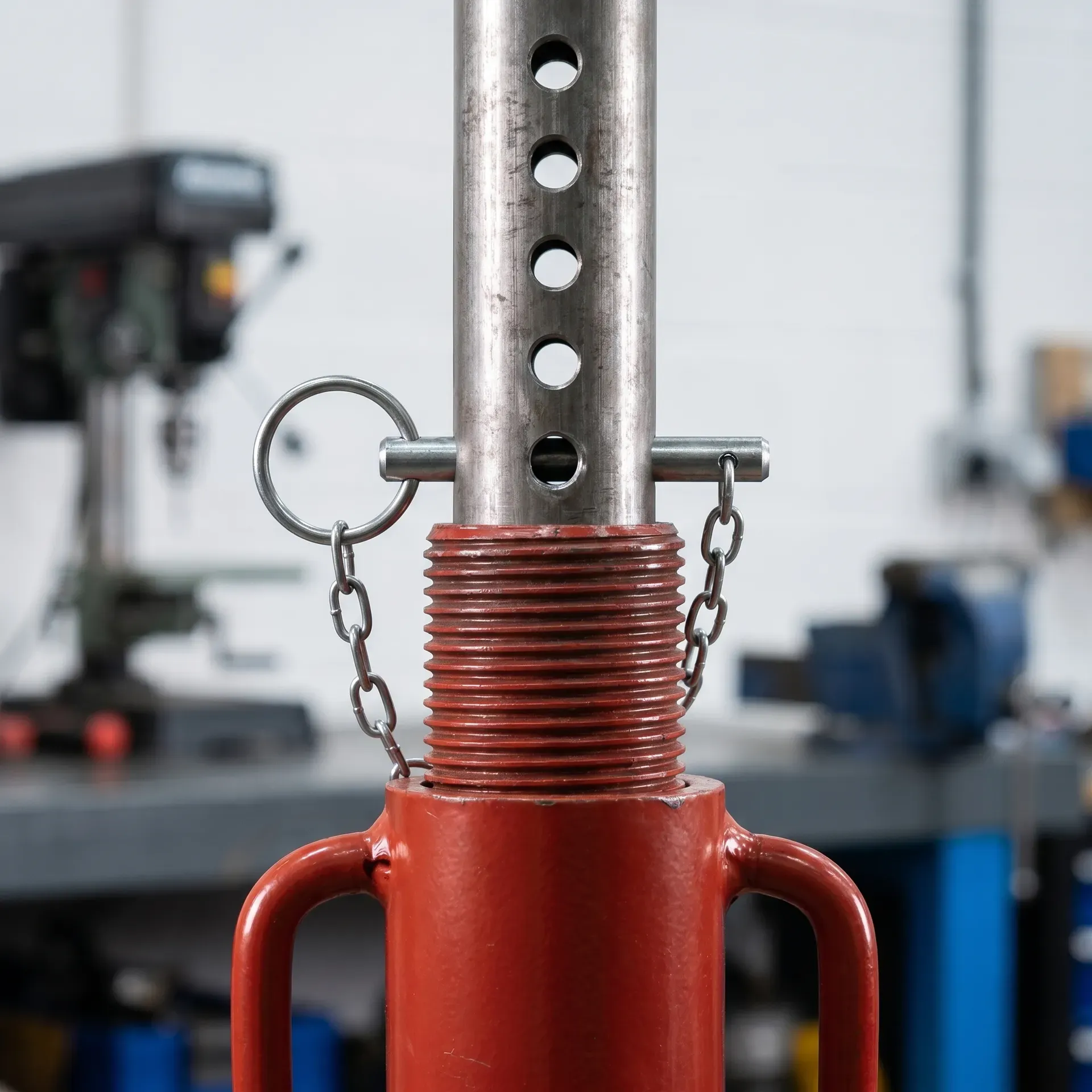 Close-up photograph of the upper section of an adjustable steel prop showing the threaded collar on the outer red-painted tube, the inner tube emerging through it with a row of pin holes, and a steel locking pin inserted through the matching holes with a chain attaching it to the prop body.