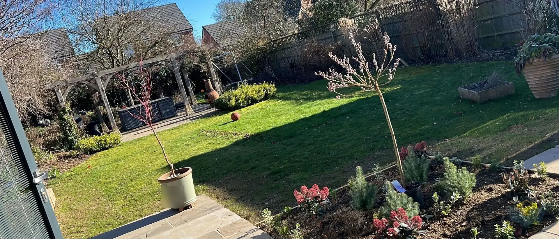 A reinstated back lawn in early spring with a paved patio and furniture to one side, newly planted garden beds to the other, and bare trees behind.