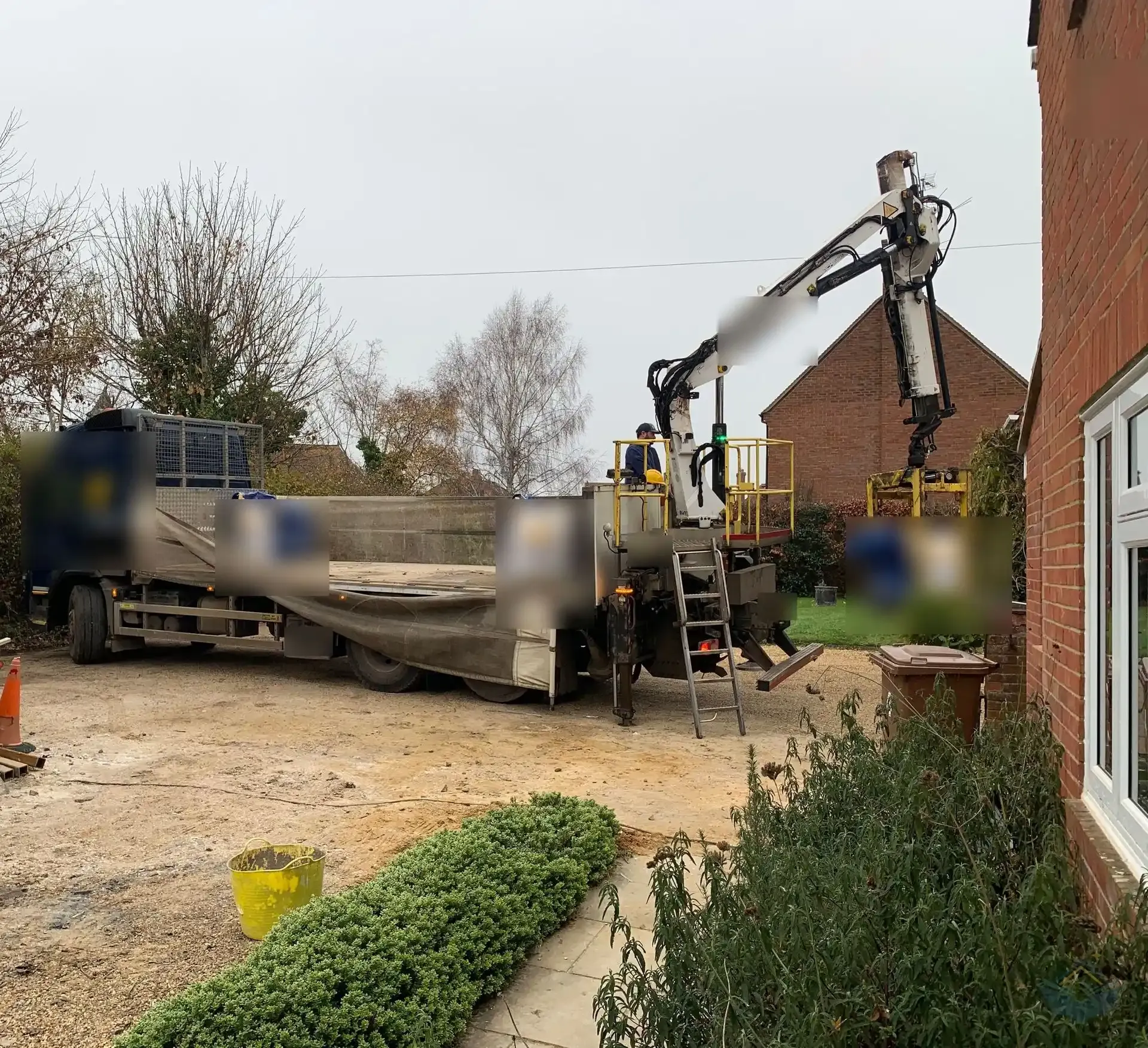 A flatbed lorry with a crane parked on a churned up gravel driveway beside a brick house, lifting large bulk bags of gravel off the bed.