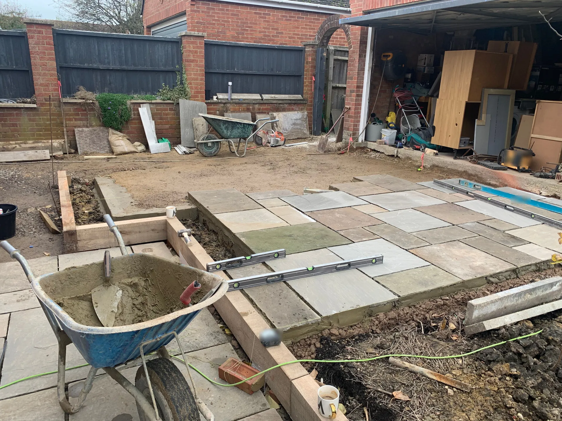 Paving slabs part laid on a mortar bed outside an open garage, two spirit levels resting across them, and a wheelbarrow of wet mortar with a trowel in the foreground.