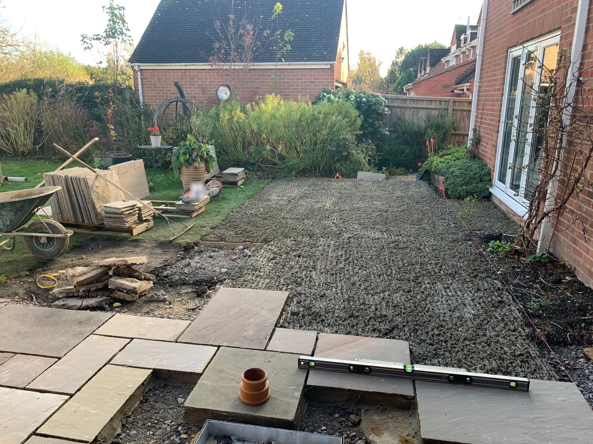 A patio under construction on a fresh mortar bed, with a part-laid run of sandstone slabs, stacks of new slabs waiting, a wheelbarrow and a spirit level.