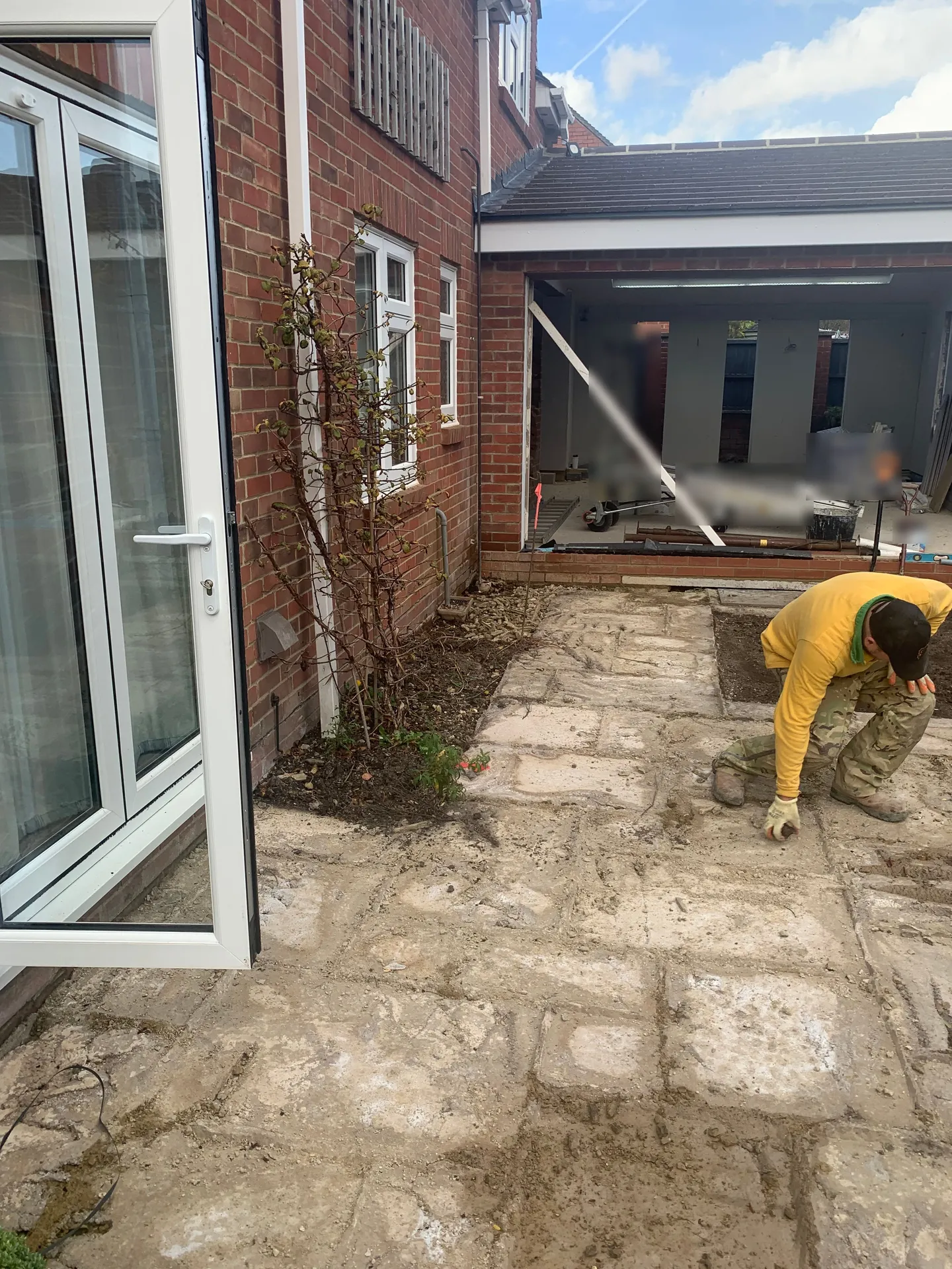 A kneeling worker lifting slabs from an old patio beside a brick house, bare earth and stacked paving under him, with the open aperture of the unfinished extension and its temporary props behind.