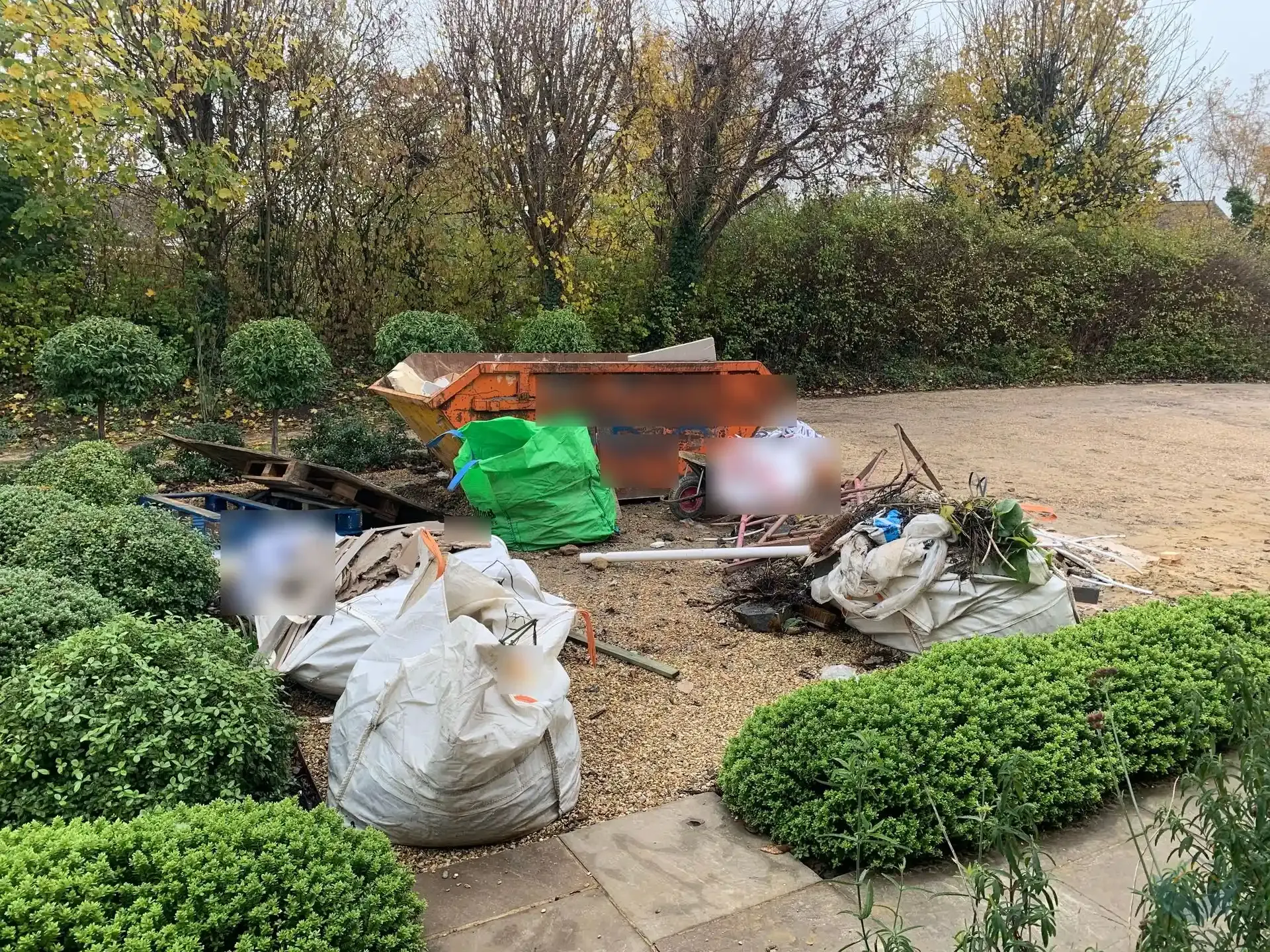 An orange skip on a gravel drive surrounded by bulk bags of waste, wooden pallets and piles of building debris, bordered by hedges and bare autumn trees.