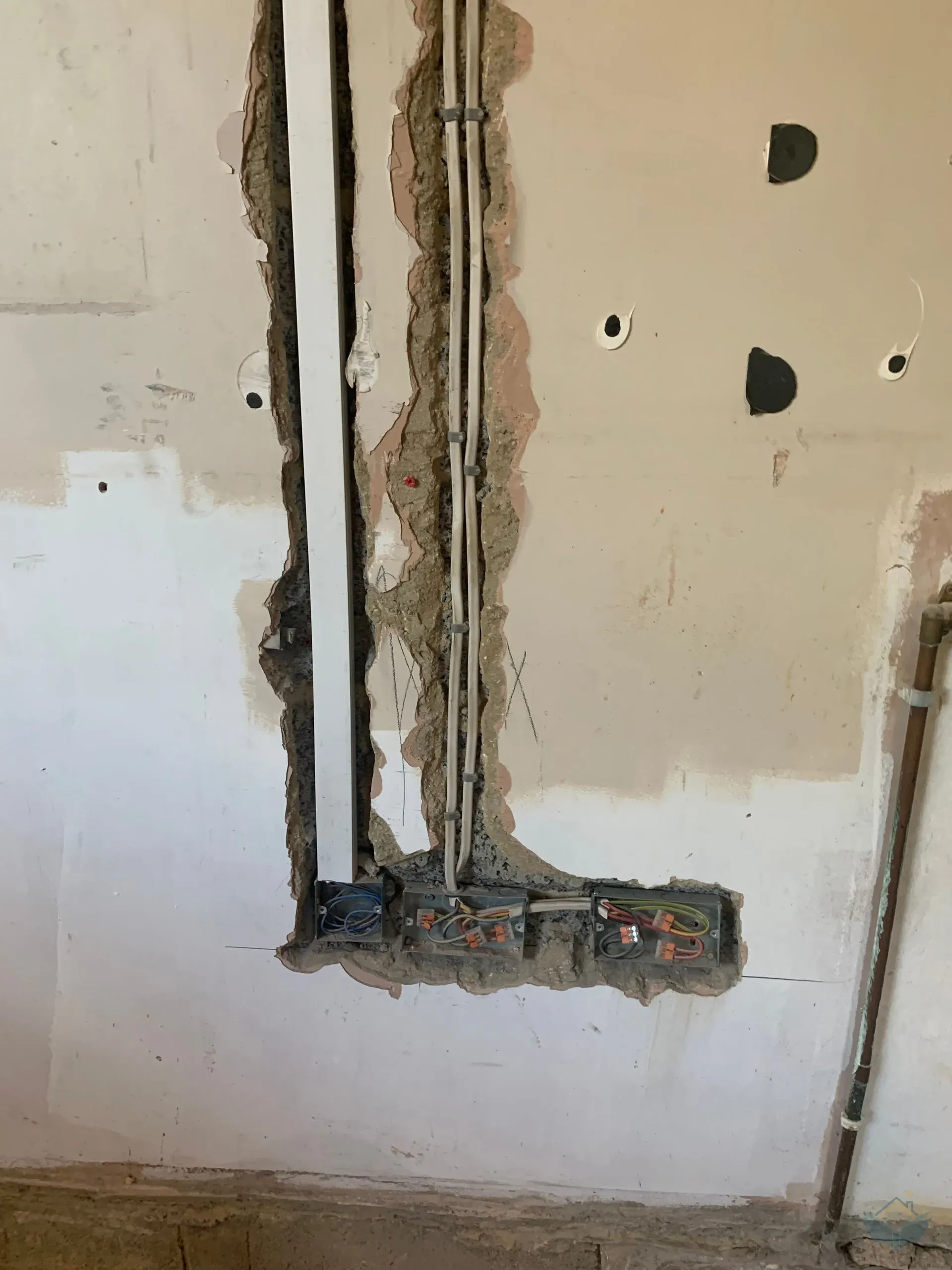 A wall with a vertical chase cut into it, two clipped cables running down inside the chase into three recessed metal back boxes at floor level containing wiring with orange connectors, and a metal pipe running vertically up the right hand side.