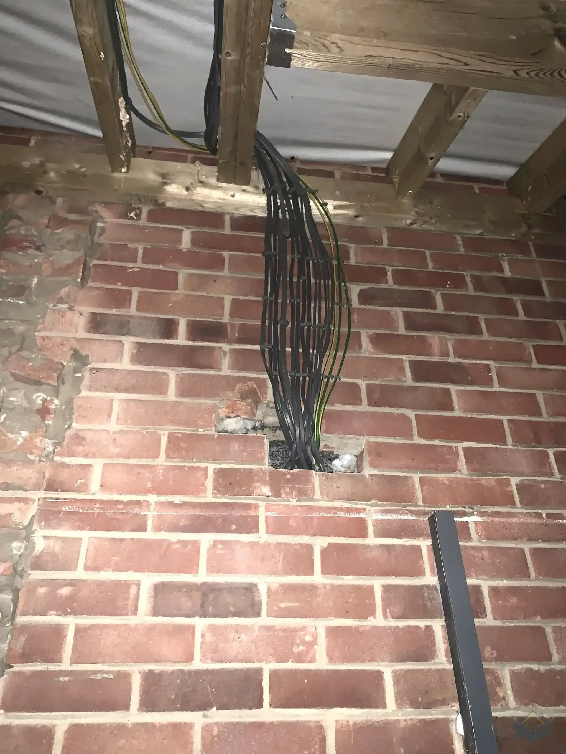Interior brick wall with an exposed timber joist ceiling above, a thick bundle of black electrical cables and green and yellow earth wires descending from the ceiling and passing through a roughly cut rectangular opening in the brickwork.