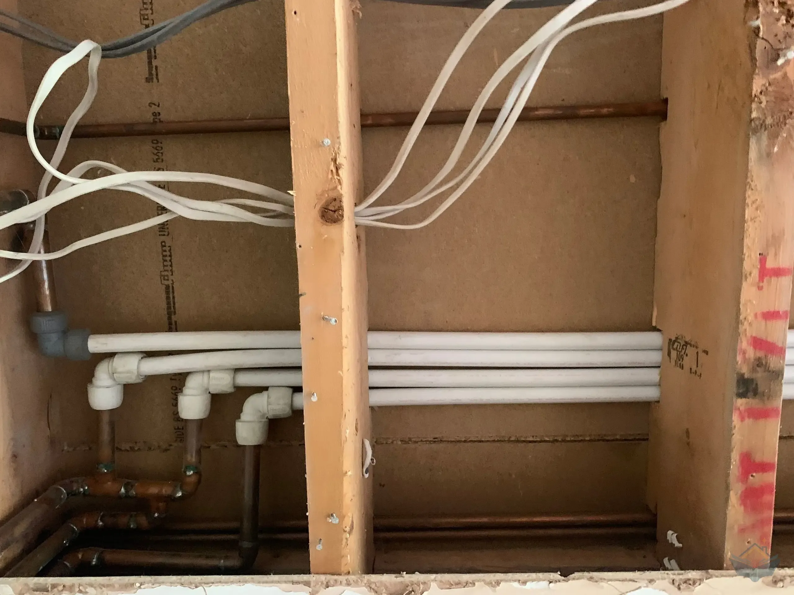 An opened wall cavity during first fix, white push-fit plastic elbows joining onto the soldered copper pipework of the original house, with electrical cables running through the timber studs above.