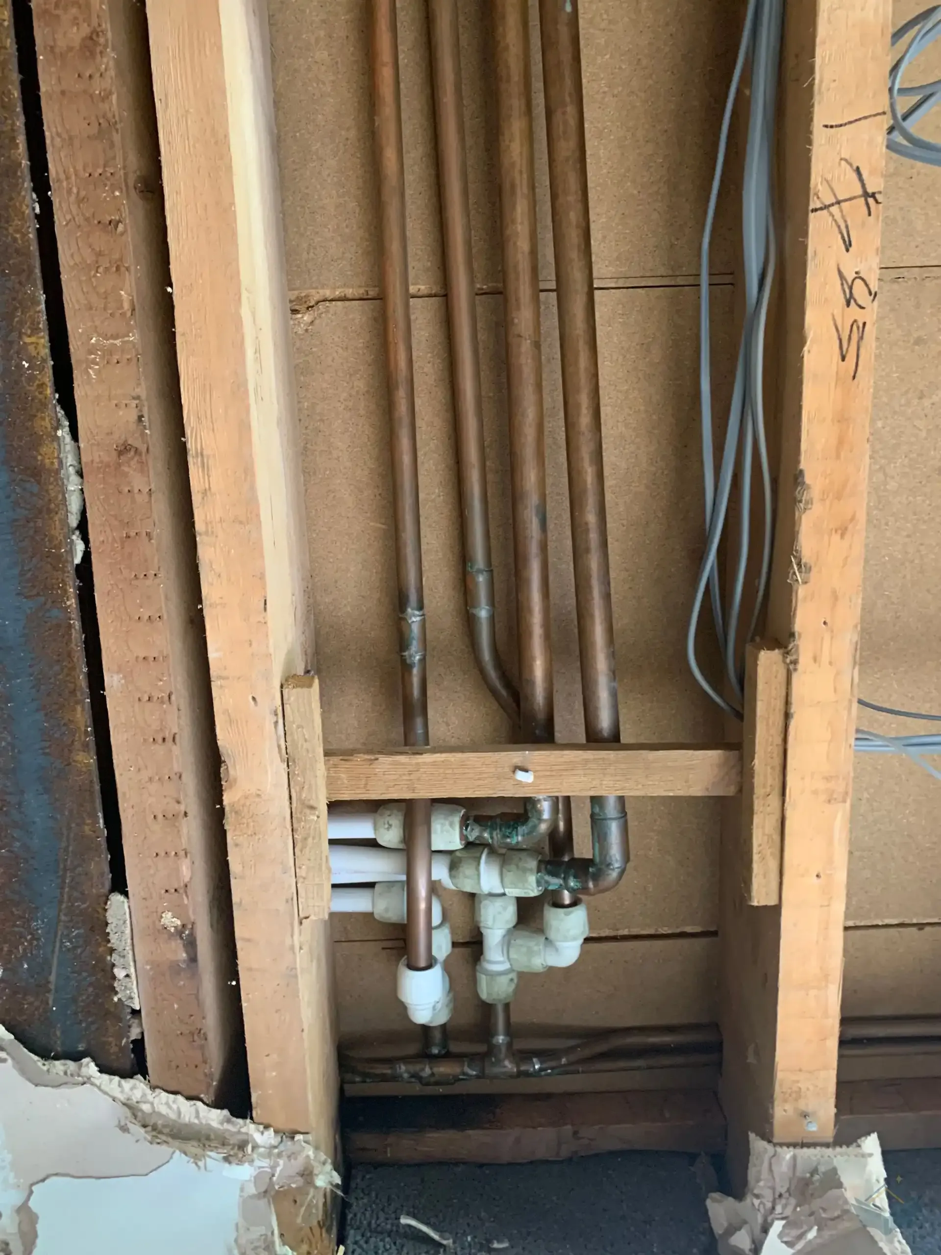 An opened wall cavity with vertical copper pipes dropping from the ceiling into white plastic push-fit fittings and horizontal copper runs below, grey electrical cables running through the timber studwork alongside.