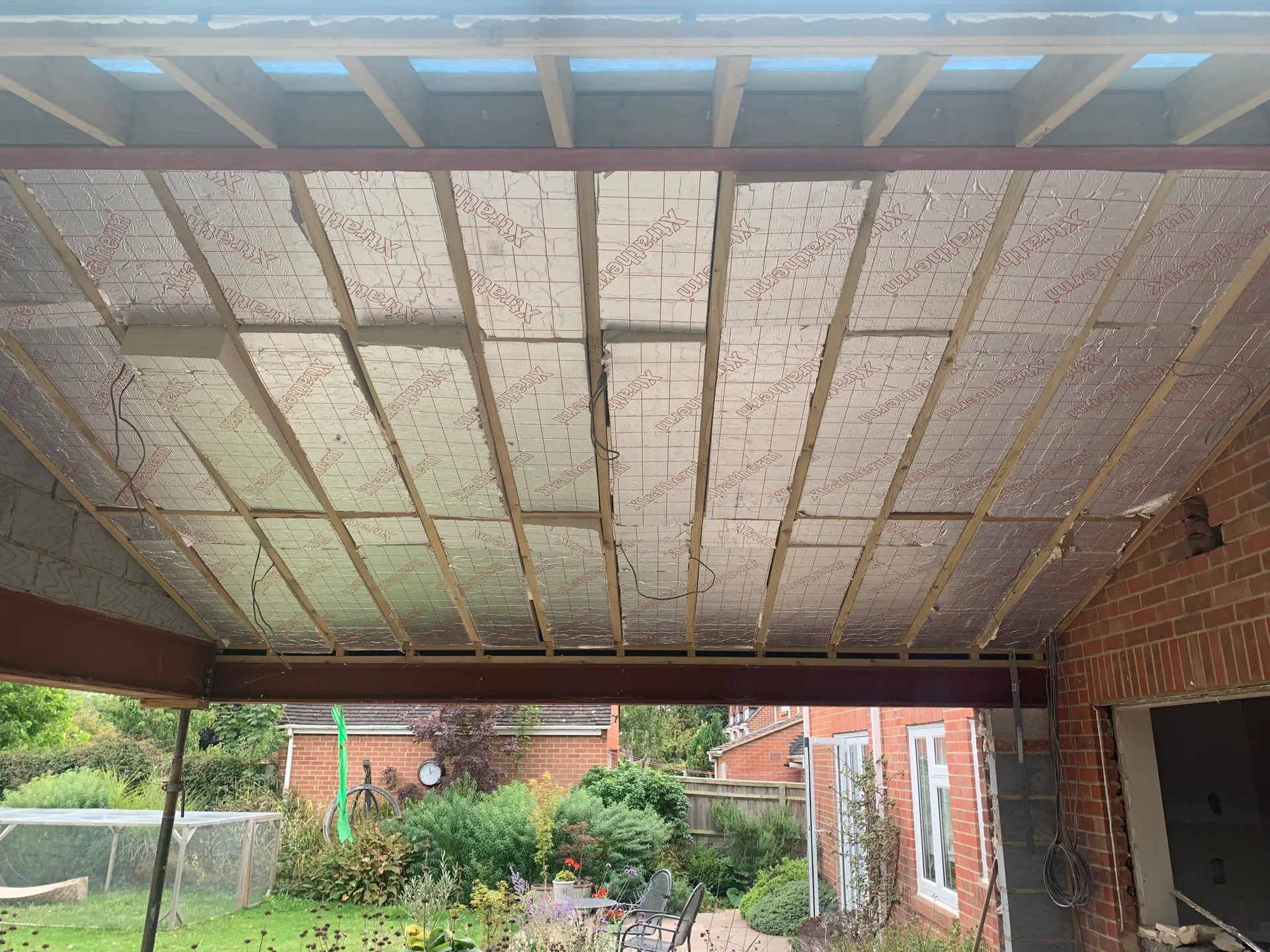 Foil faced PIR insulation boards fitted between deep timber roof rafters, seen from inside an extension, with a painted steel beam below and a garden visible through the open wall beyond.