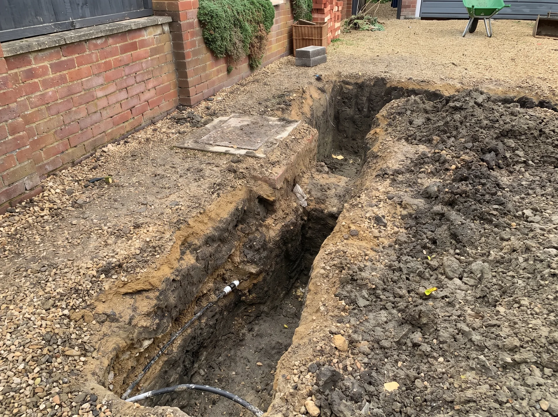 An excavated trench showing a drain run passing under the line of the new extension wall towards a manhole cover on the driveway.