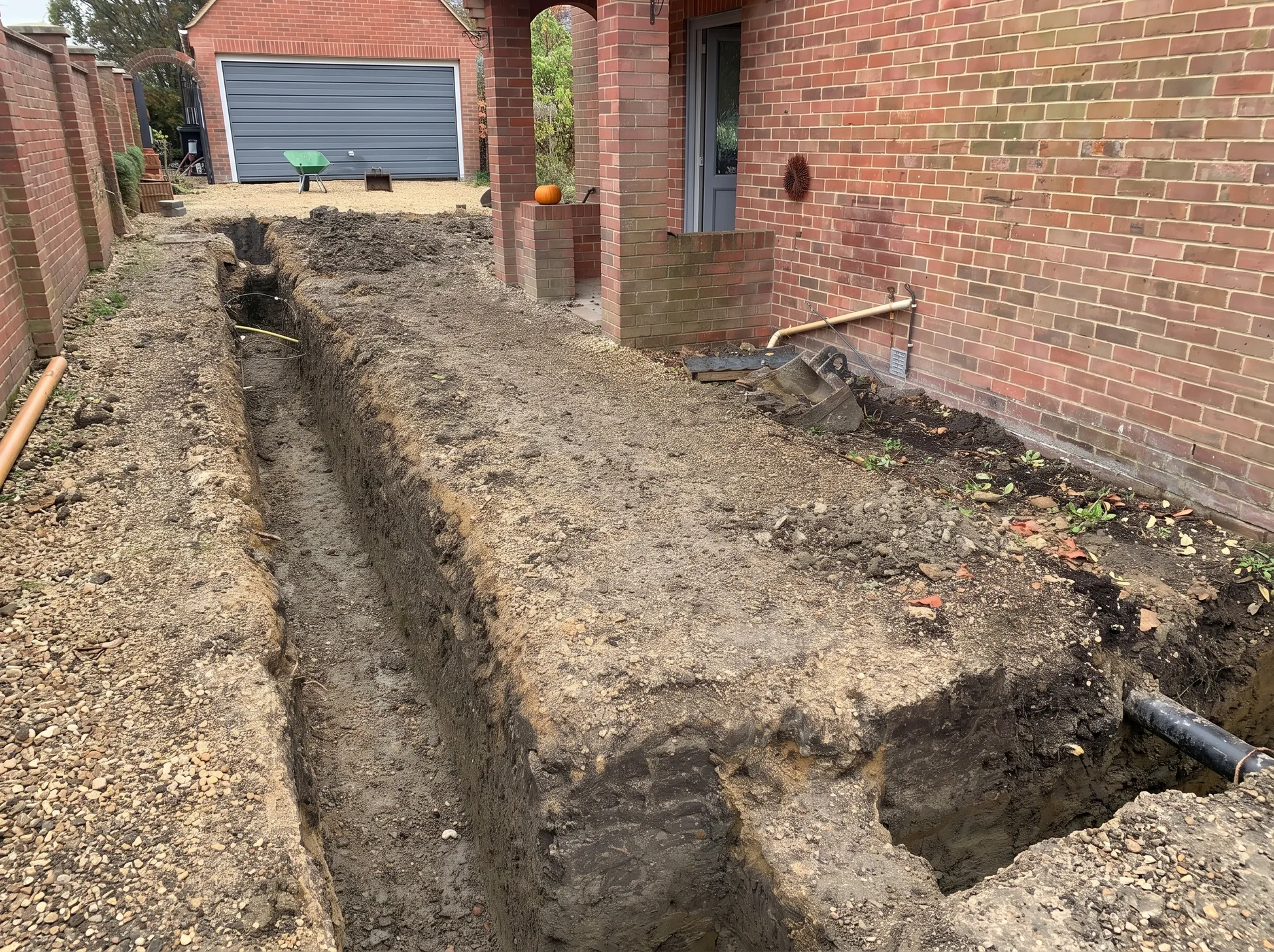 A deep foundation trench dug around the perimeter of an extension footprint, with exposed pipework against a brick wall, spoil heaped either side and a manhole cover in the foreground.