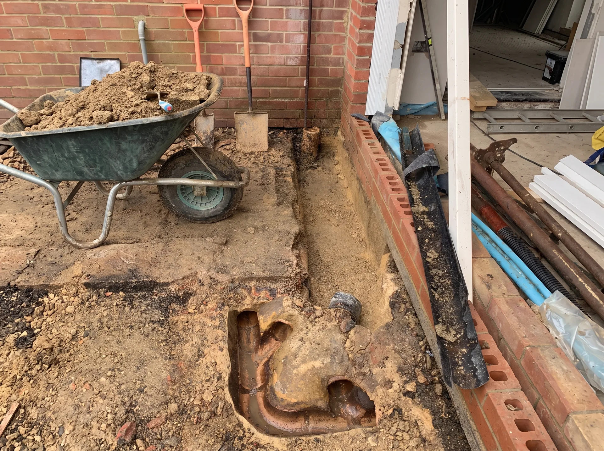An excavated trench alongside a brick wall exposing old ceramic drainage pipes with several branch connections, partly filled with water.