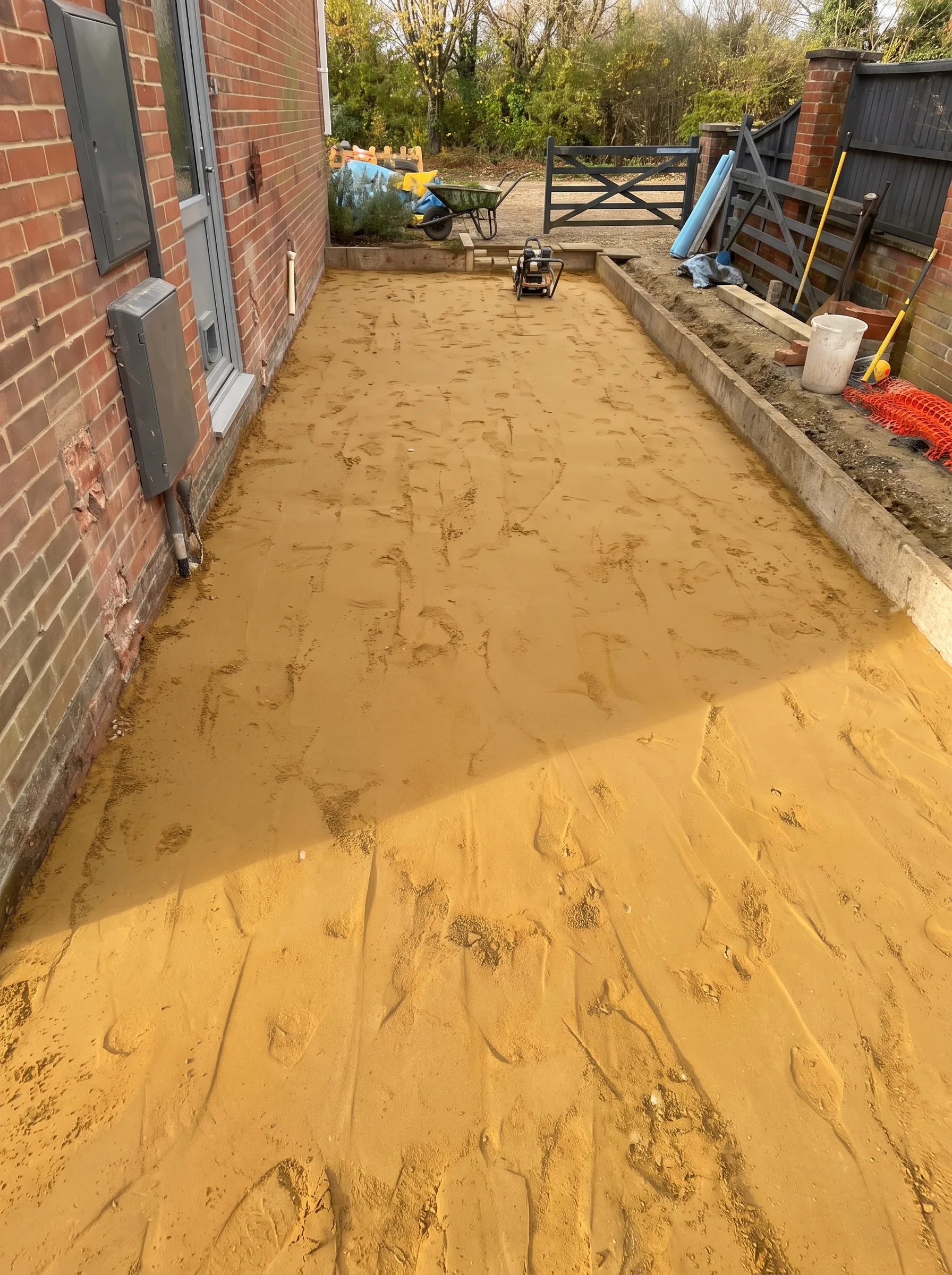 A rectangular area of compacted hardcore covered in a smooth layer of sand, bordered by blockwork, with a plate compactor and tools nearby.