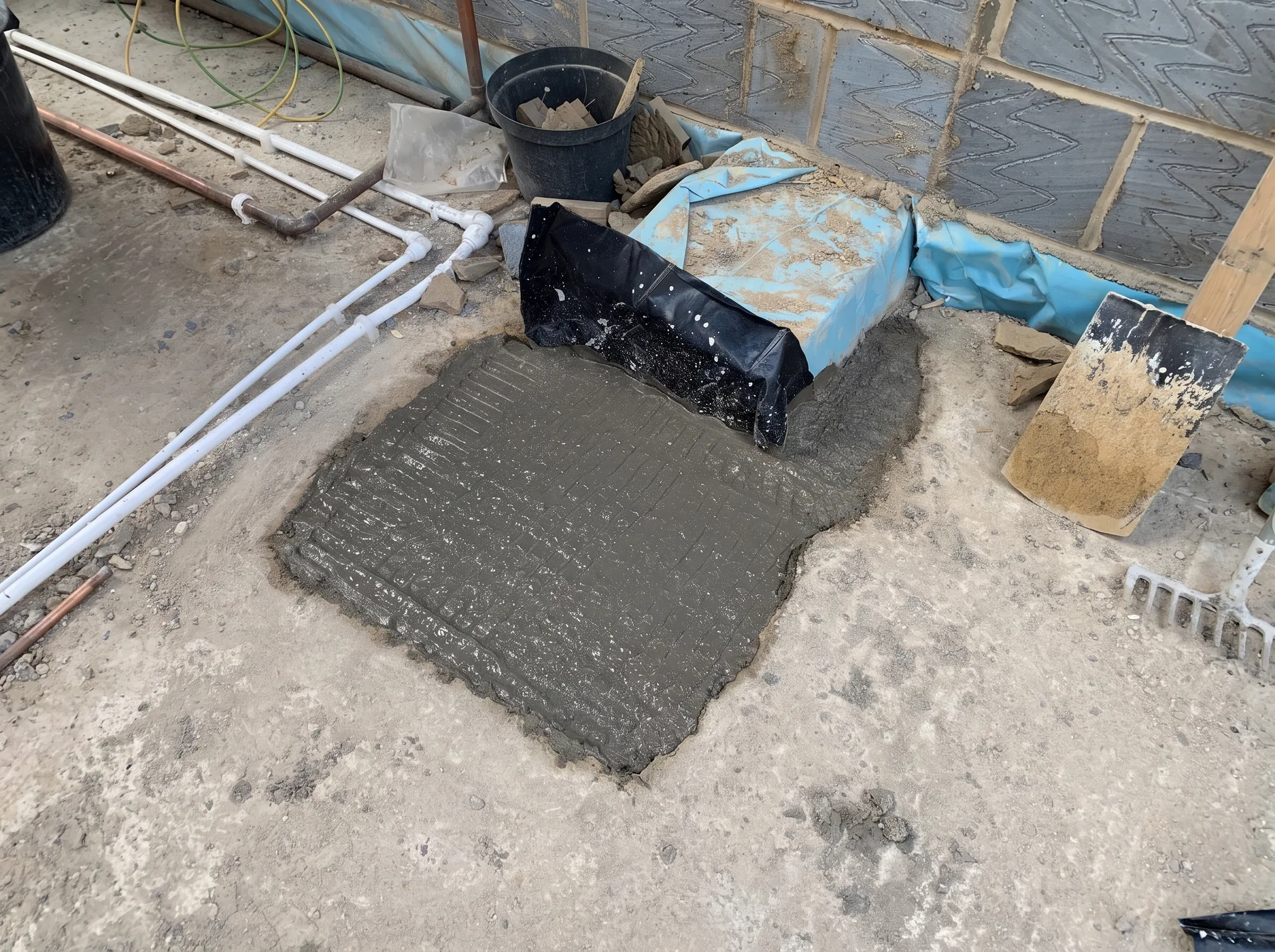 Freshly poured concrete filling a rectangular excavation cut through an existing floor slab, with overlapping damp proof membrane and pipework around it.