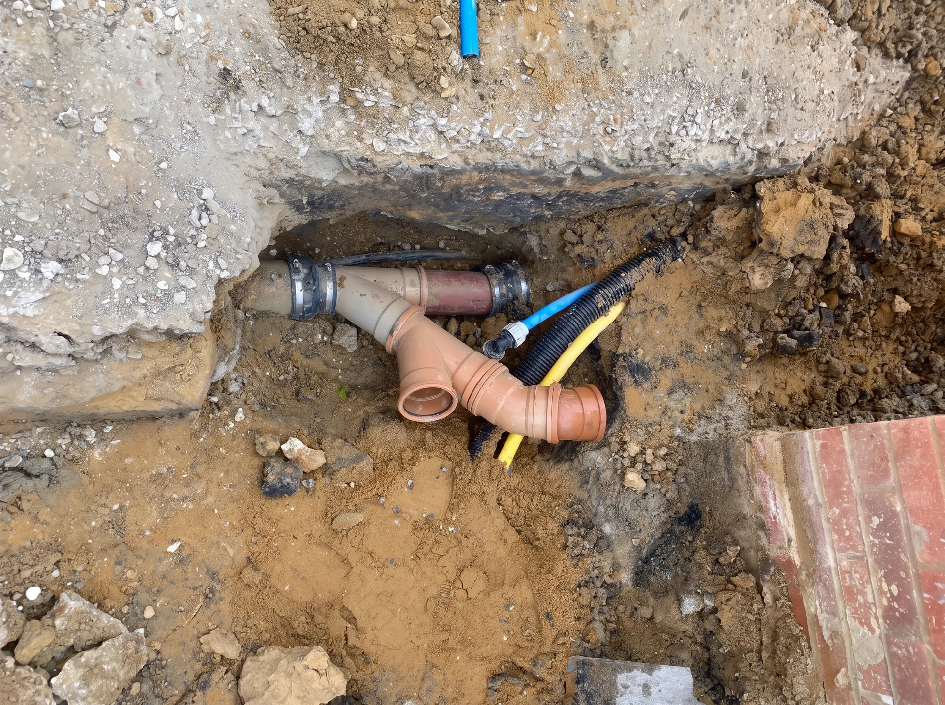 Close-up of a shared trench containing gas, electricity and water services alongside an old ceramic sewer pipe repaired with a plastic section and flexible couplings.