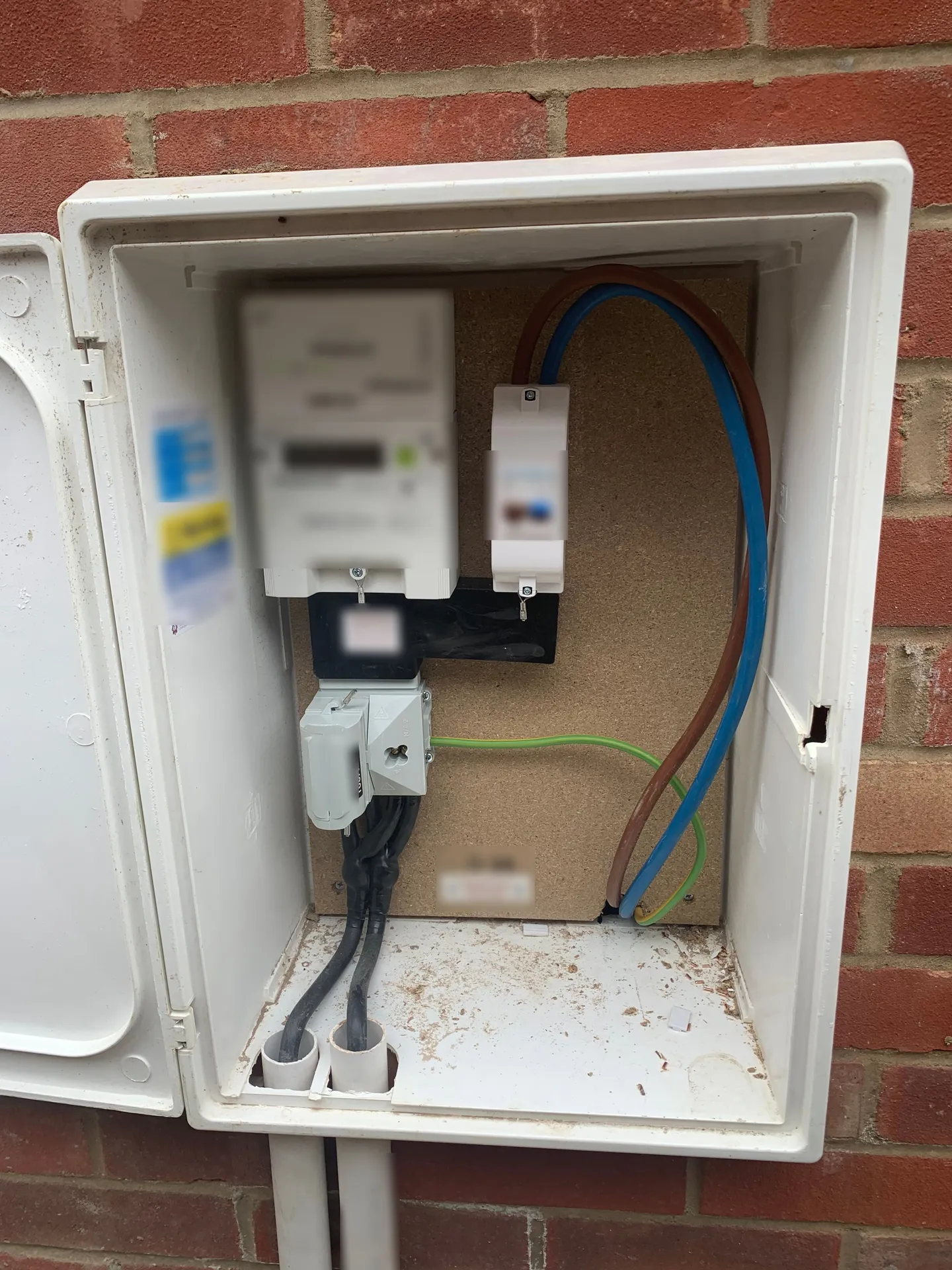 An open white meter cabinet set into a red brick wall, an electricity meter and control gear mounted on a wooden backboard inside it, supply cables entering from below through white conduit.
