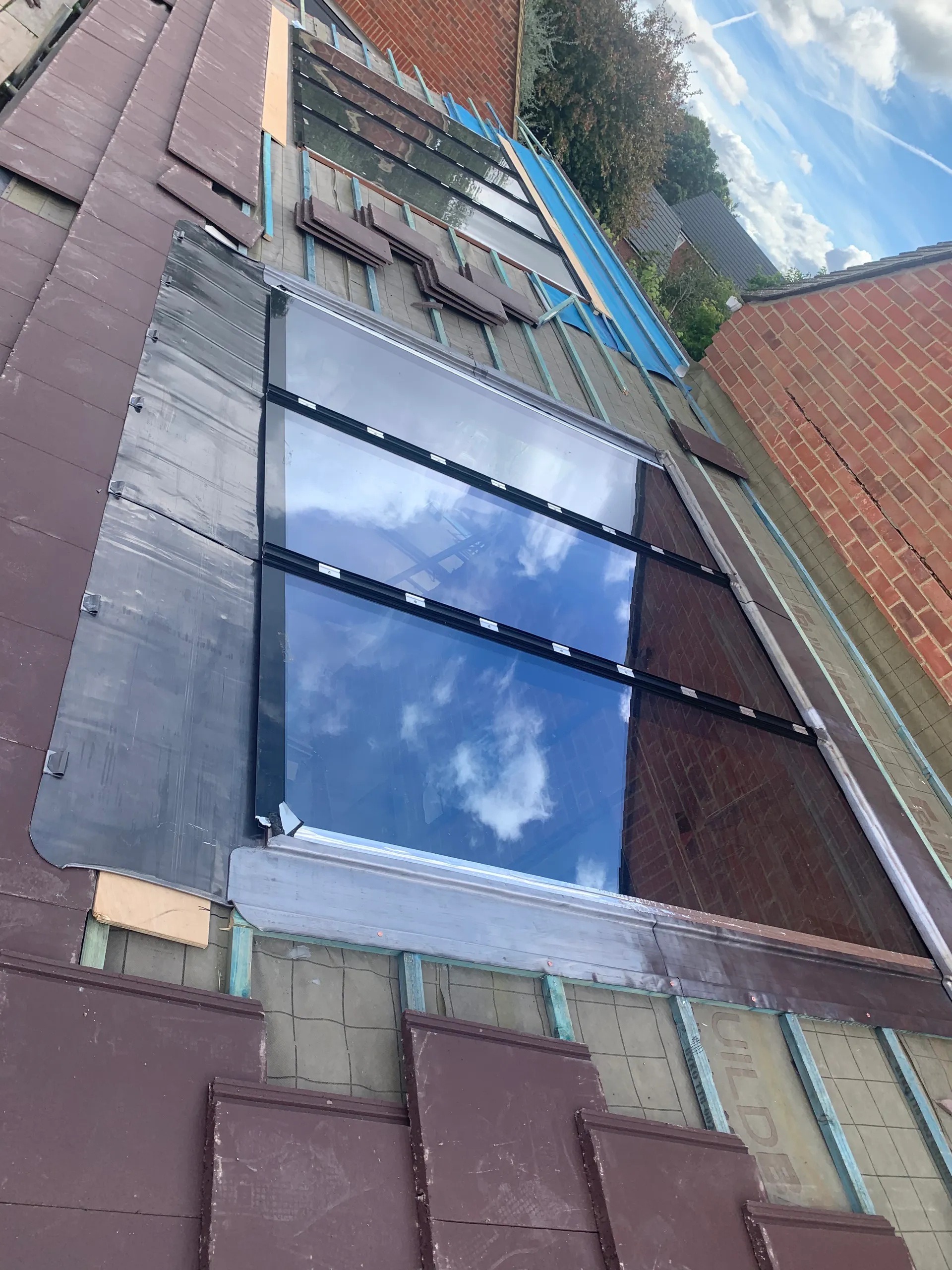 A pitched roof under construction with a large multi pane roof window leaded around its edges, a second bare window beside it, clay tiles partly laid over battens and membrane.