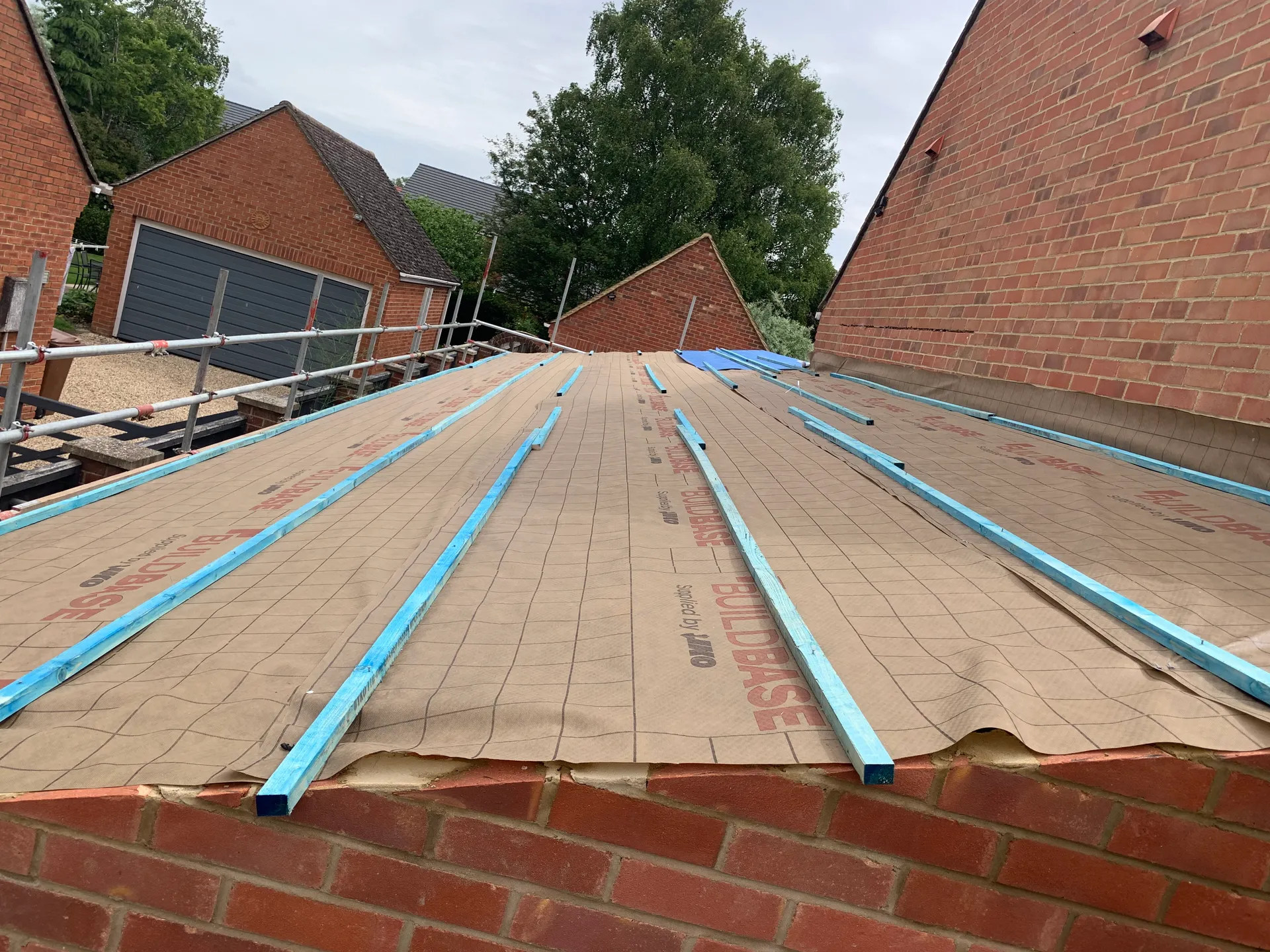 A low pitched roof covered in breathable membrane with parallel timber battens laid over it, brick walls and scaffolding around it under an overcast sky.