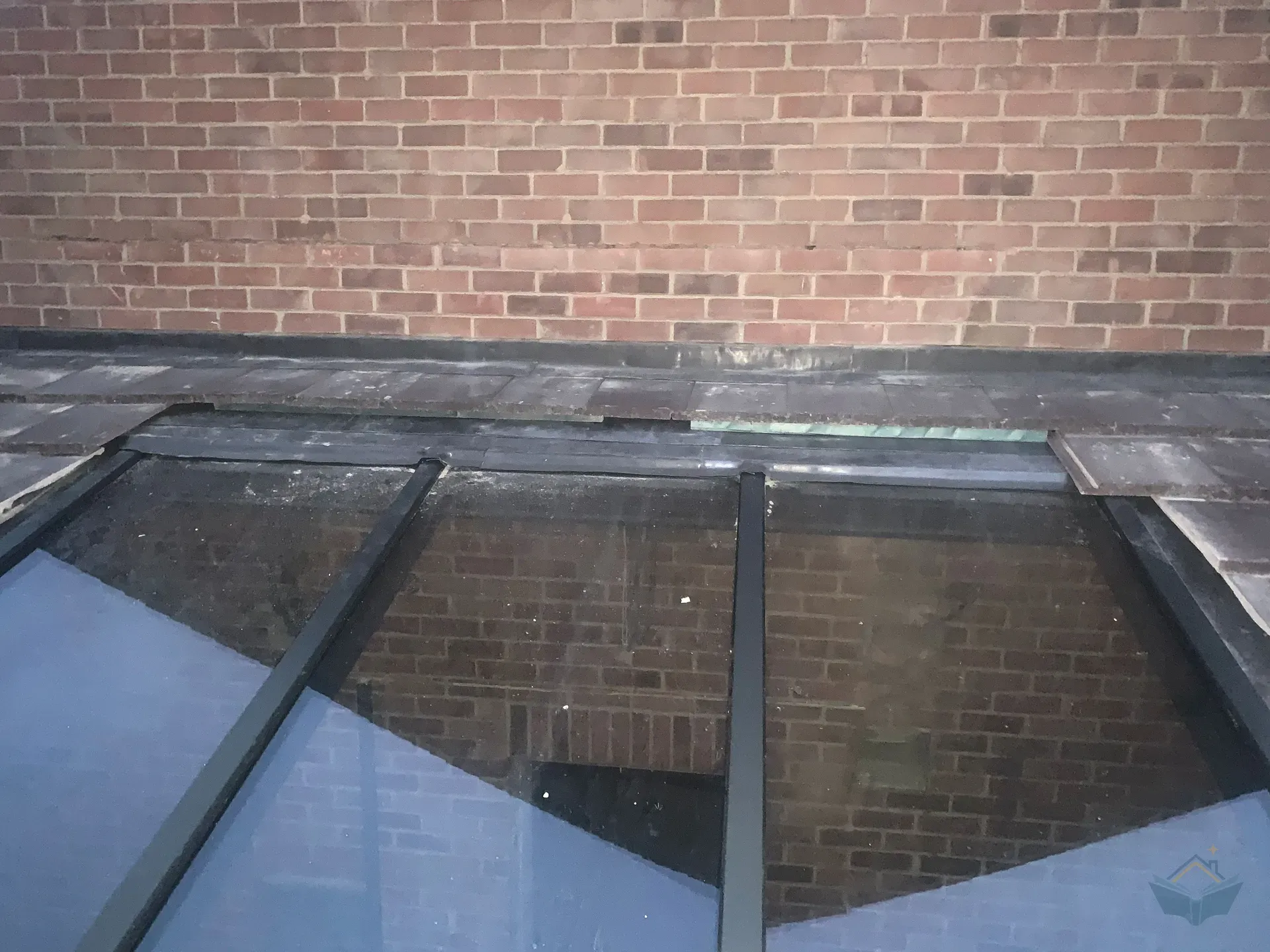 Exterior view along a tiled roof towards a large roof window beneath a brick wall, with an open gap between the cut tile course and the top of the window frame.