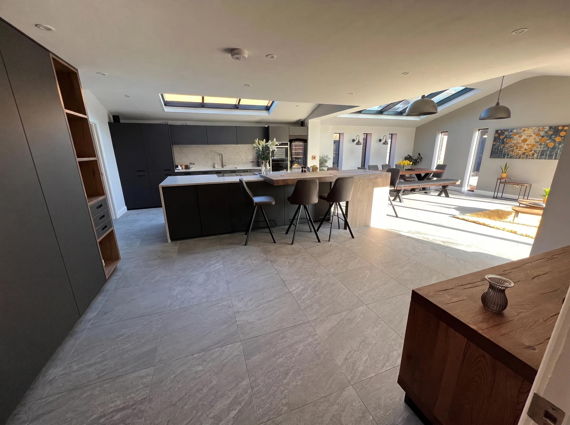 Finished open-plan kitchen with dark grey cabinetry, a large island with a wooden breakfast bar, grey tiled floor, multiple skylights, dining table and bench seating