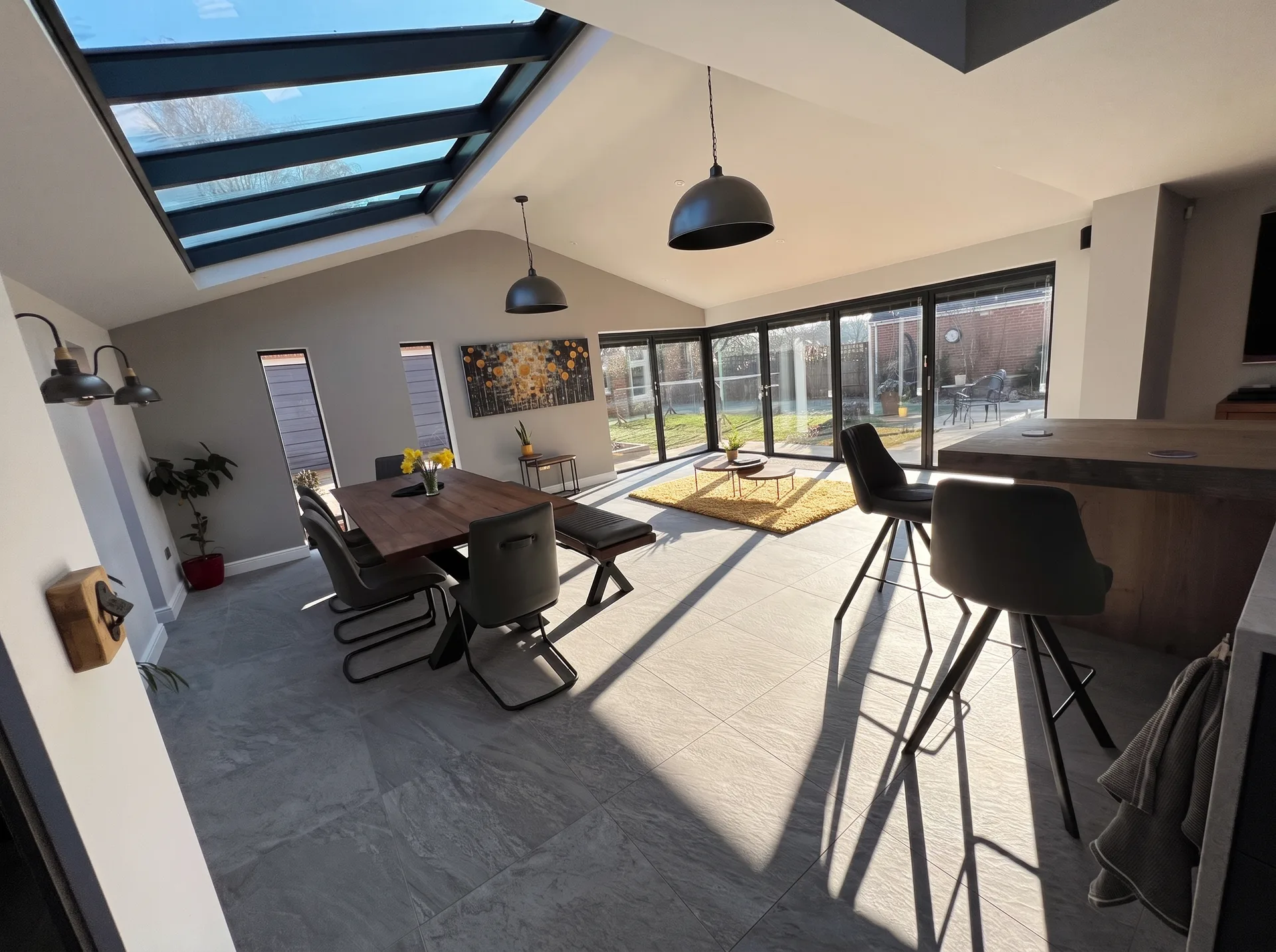 Completed open-plan living and dining space with grey tiled floor, dark furniture and pendant lighting, lit by a large roof window and bifold doors onto the garden