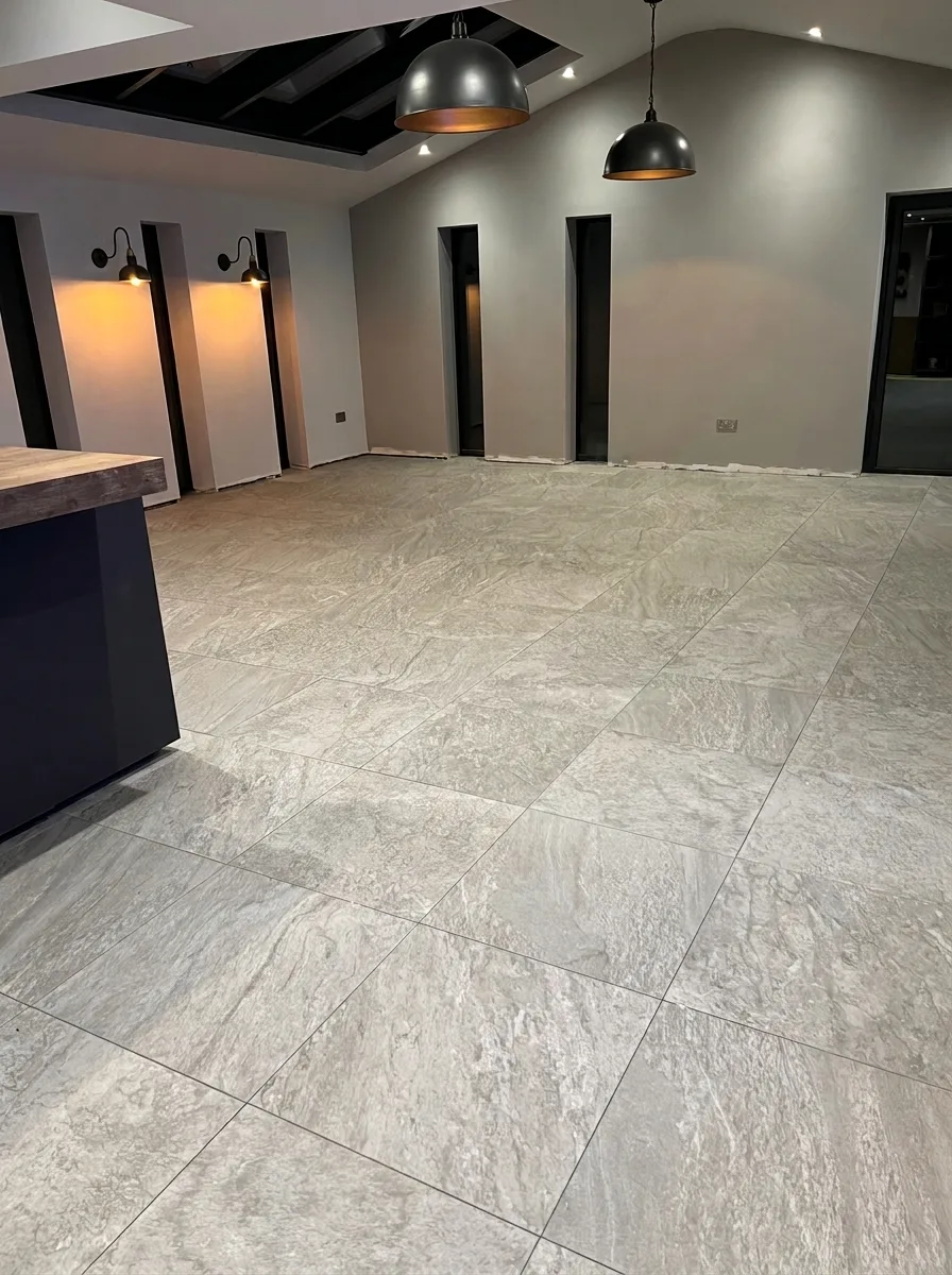 Newly laid light grey floor tiles across a kitchen with pale walls, a sloped ceiling, a large skylight and black pendant lights above a dark island