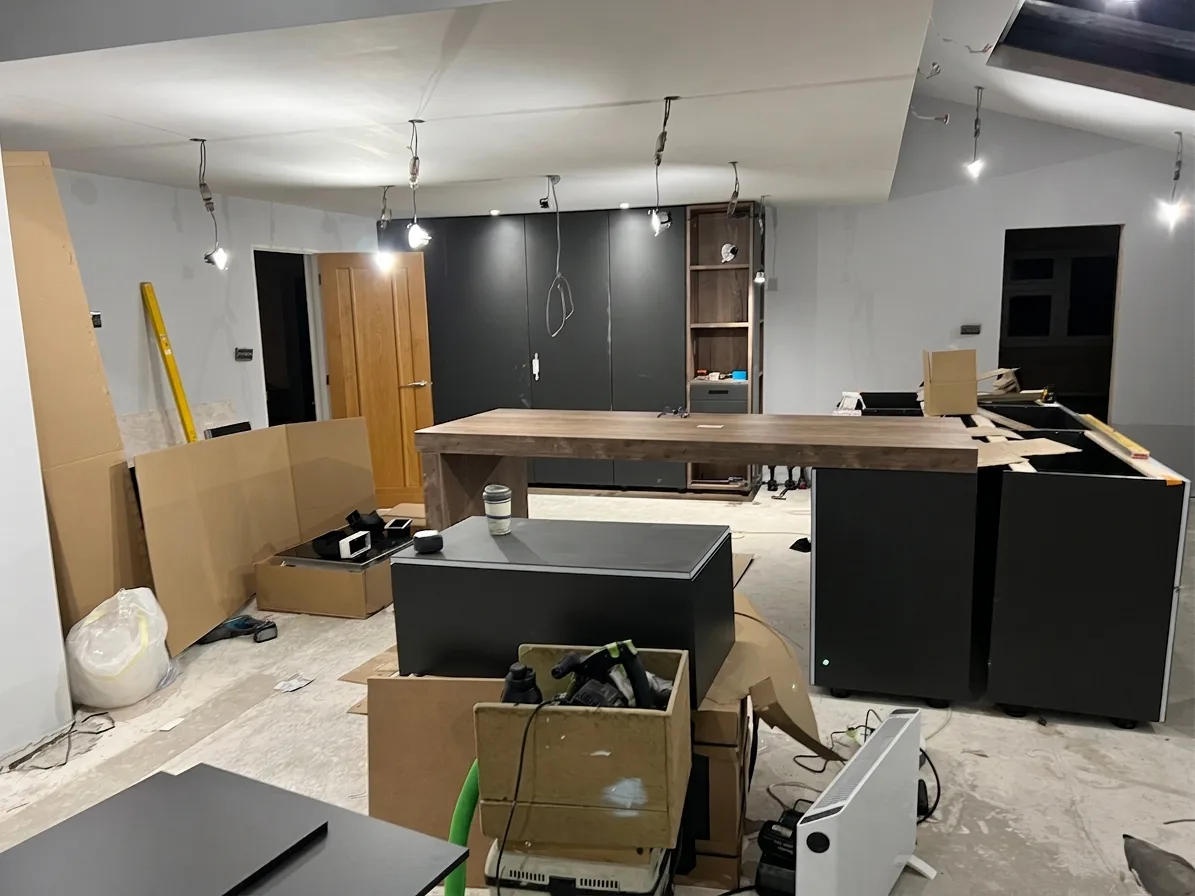 Newly installed dark floor-to-ceiling kitchen cabinets along a back wall with a large island carcass in the centre, surrounded by packaging and tools on a bare floor