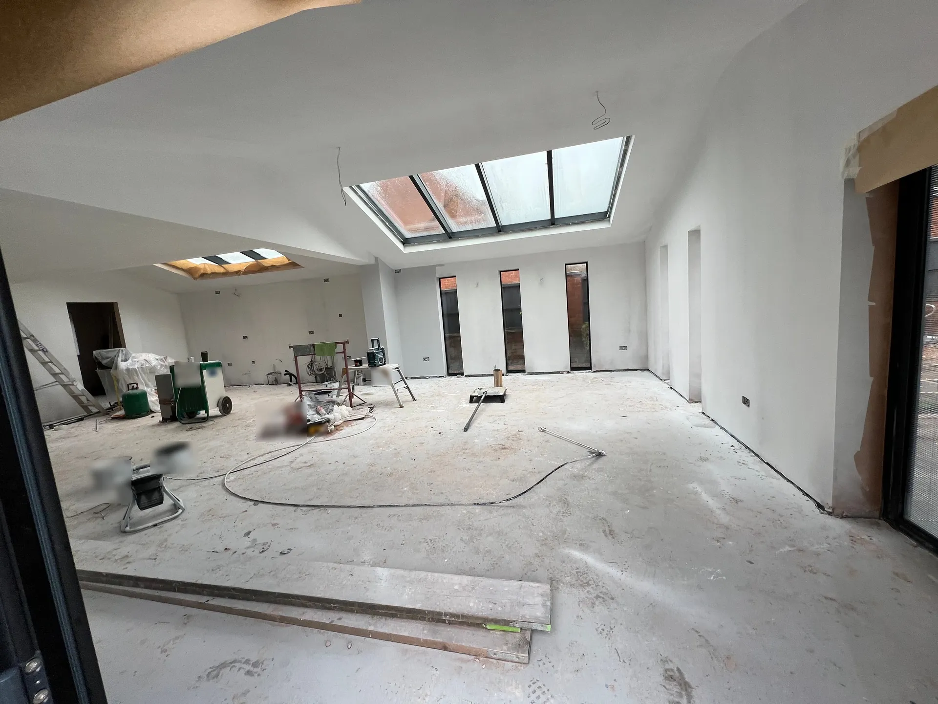 Large open-plan room with freshly plastered walls and ceiling carrying a thin white first coat of paint, bare screed floor covered in dust and footprints, with a dehumidifier, a ladder and planks standing about
