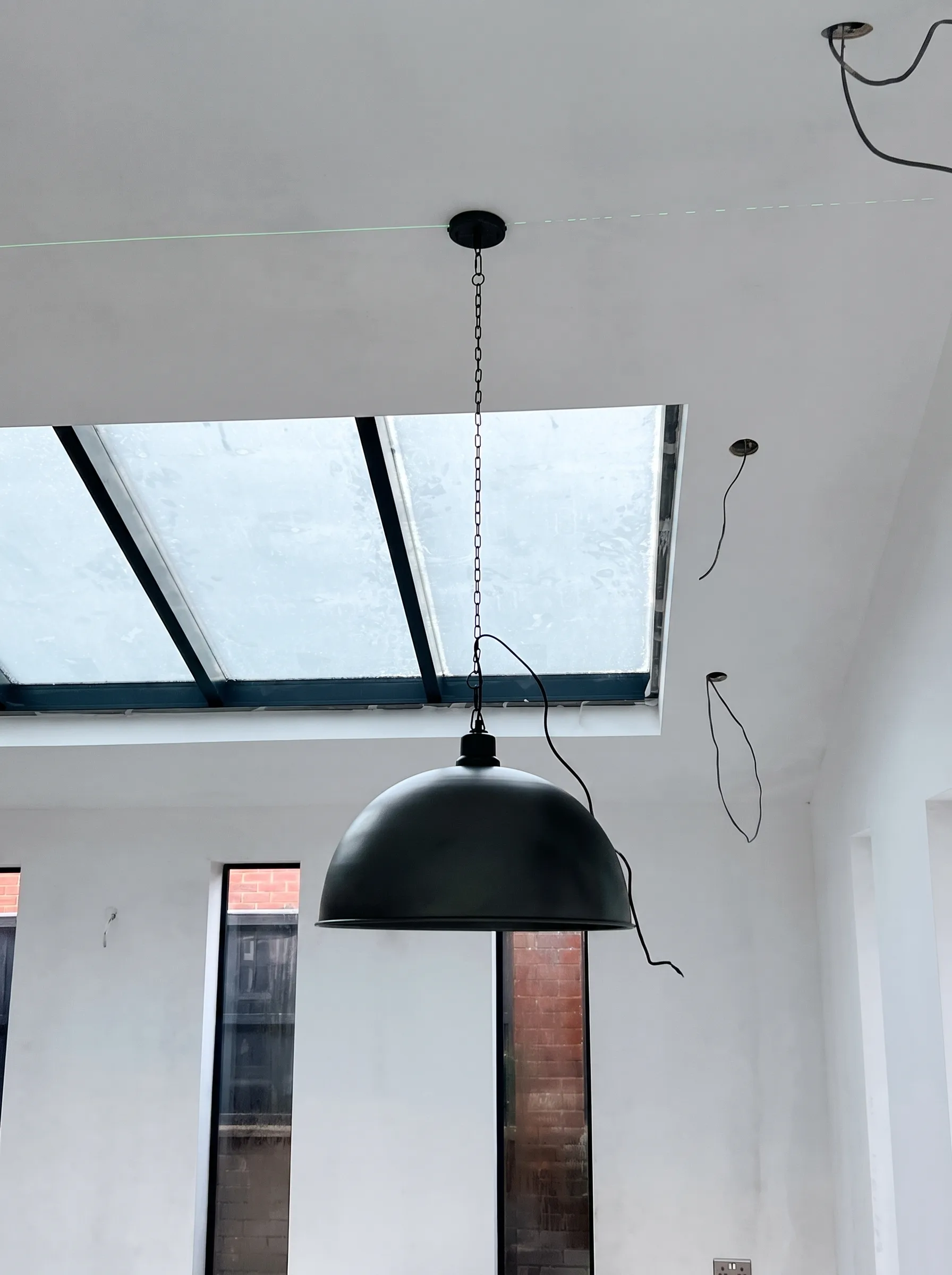 Large dark dome pendant hanging from a chain in an unfinished room, with a green laser line projected across the ceiling and circular openings for further lights