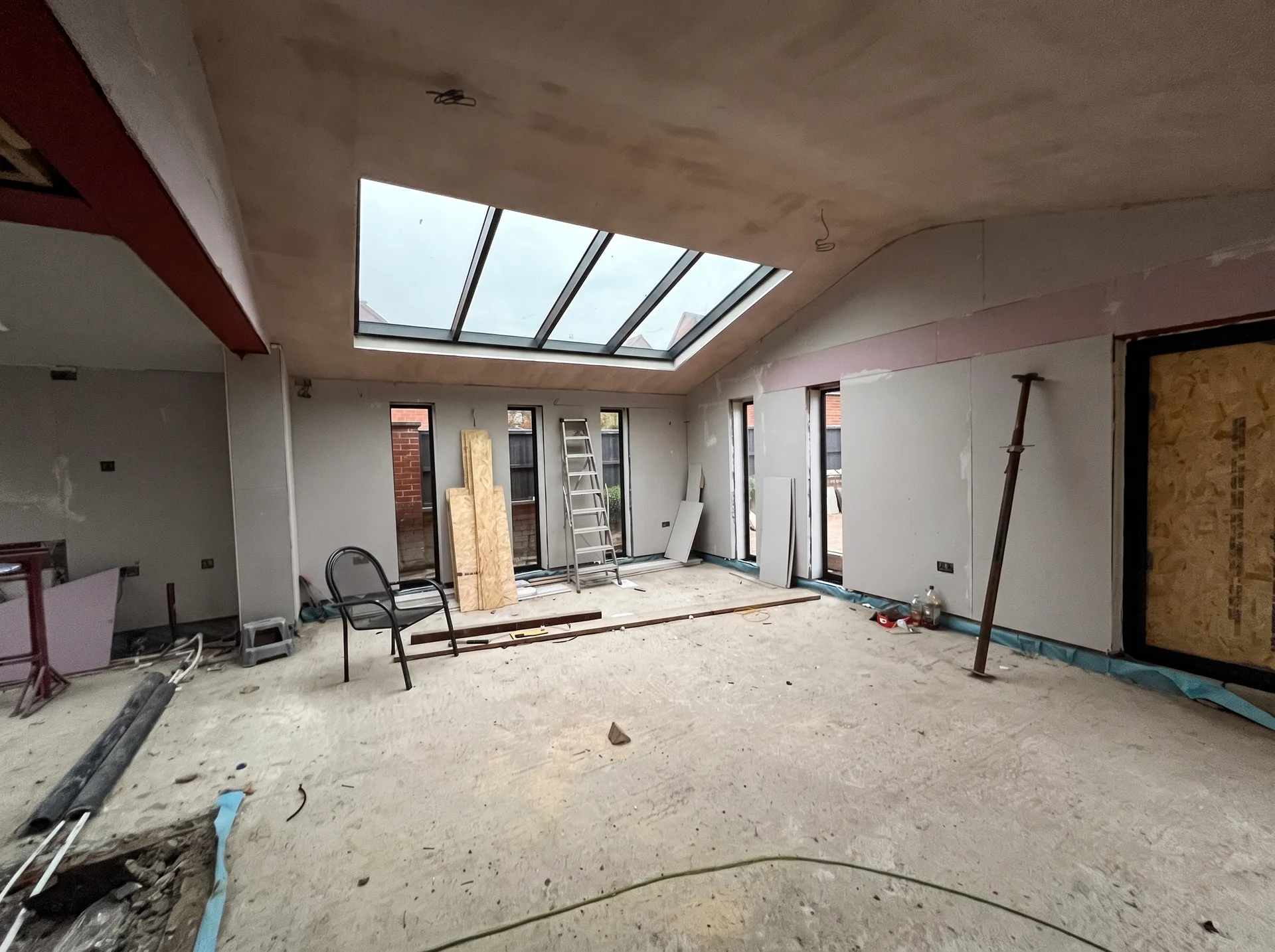 Freshly plastered apex wall at the end of a vaulted kitchen with full-height glazing installed and exposed steelwork at the rear corner