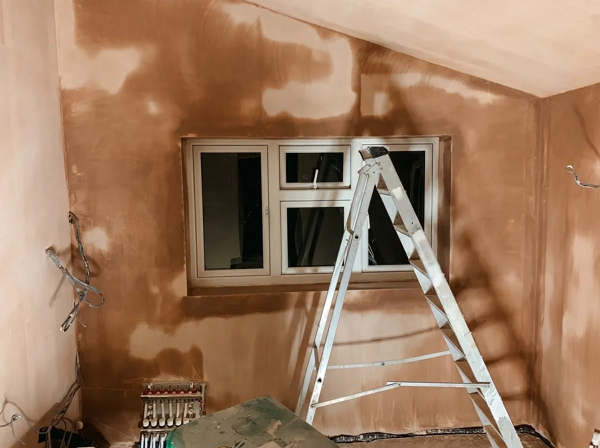 Small utility room with freshly applied wet plaster on every wall, a window in place, and an underfloor heating manifold mounted low on the left-hand wall