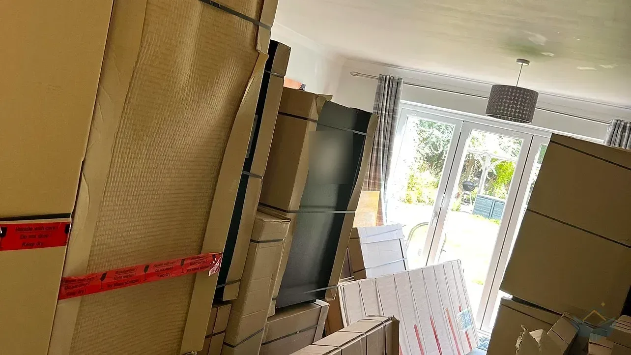 A living room filled floor to ceiling with the delivered kitchen, tall flat cardboard cabinet packs leaning against the walls and a boxed fridge freezer stacked in front of the patio doors.