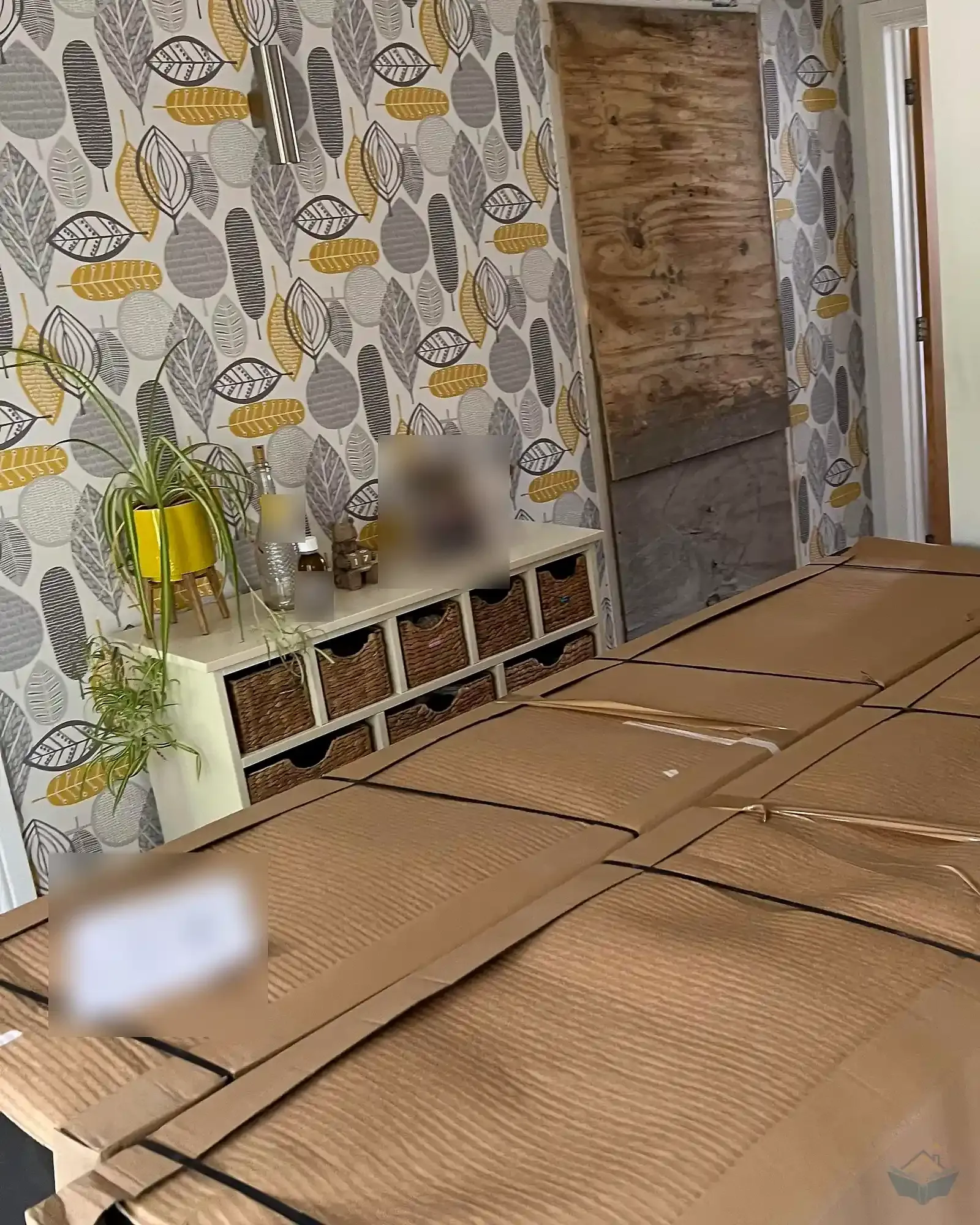 A domestic hallway with large flat cardboard packs of kitchen cabinet doors laid across the floor and stacked against the wall, beside a boarded up doorway covered with plywood.