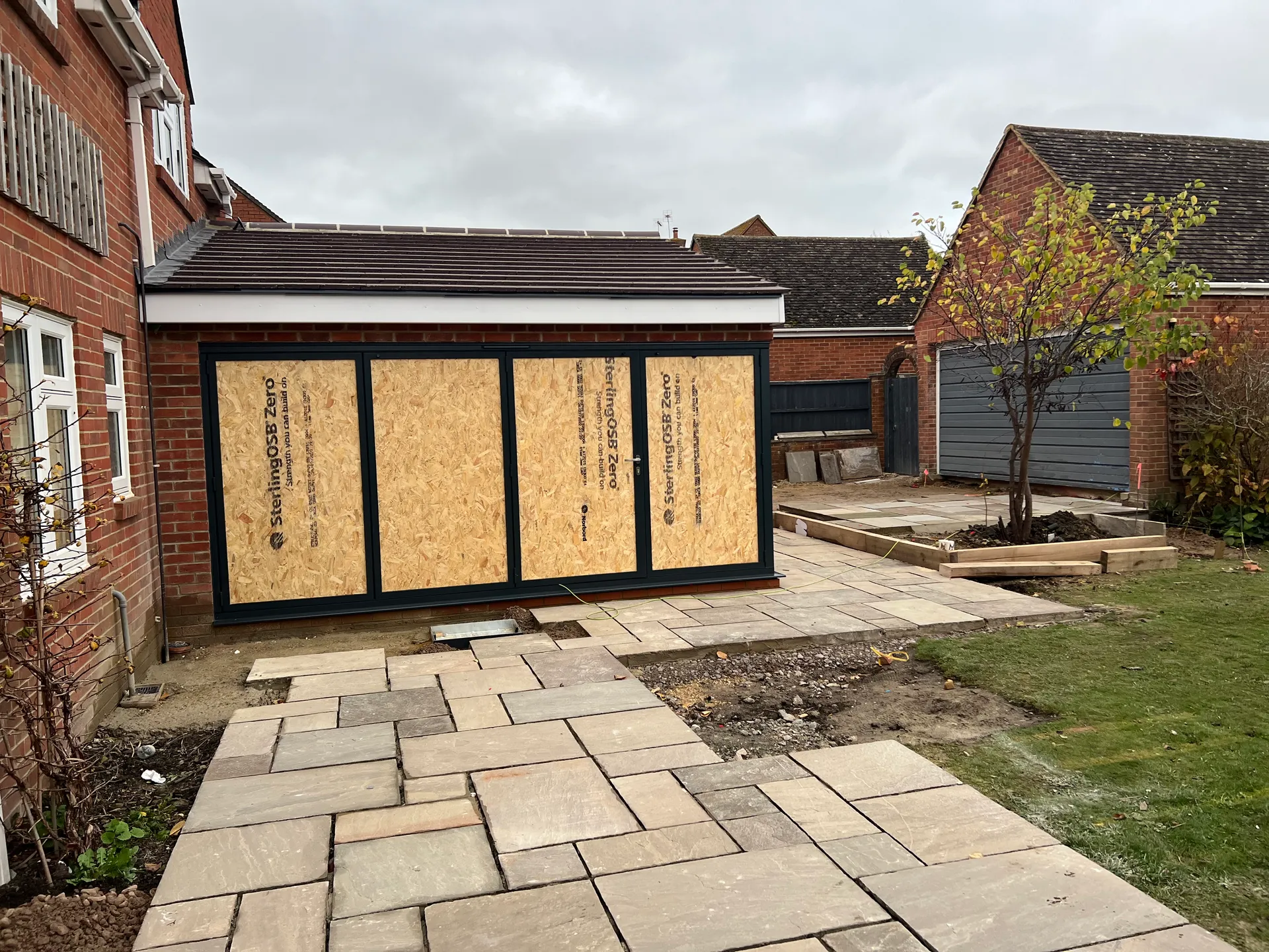 Rear of a newly built brick extension with a tiled roof, its large door openings temporarily covered with plain boards, part laid paving and disturbed earth in front of it.
