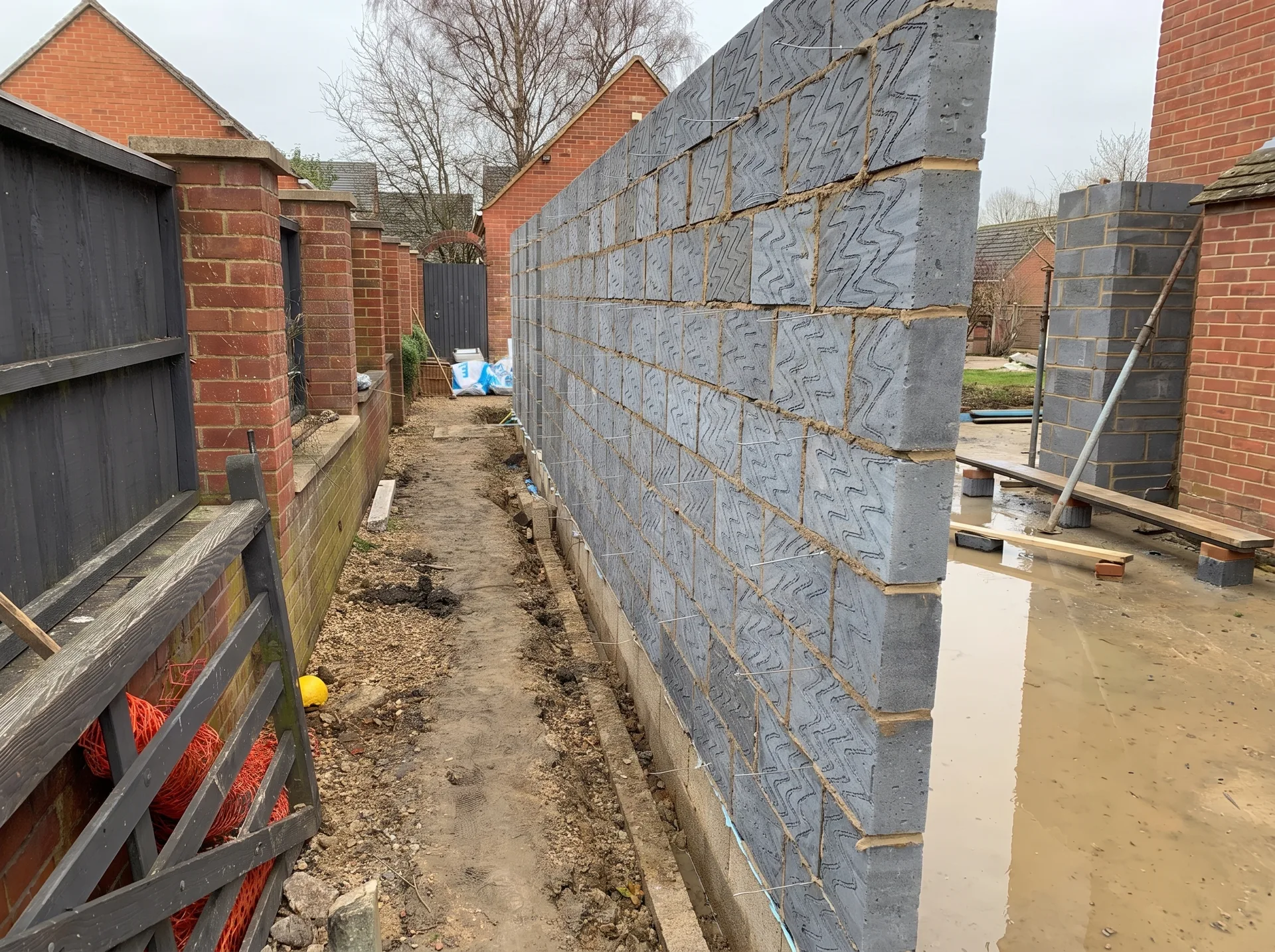 A long new grey blockwork wall running down a narrow side passage, wall ties projecting from the cavity face, muddy ground with standing water in front of it.