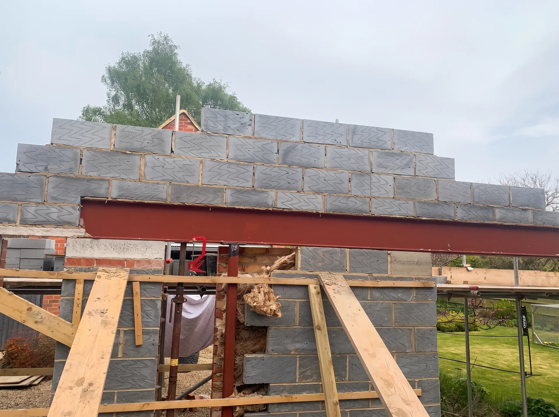 Partially built blockwork gable with a painted steel beam installed horizontally across an opening, supported on temporary timber bracing and metal props.