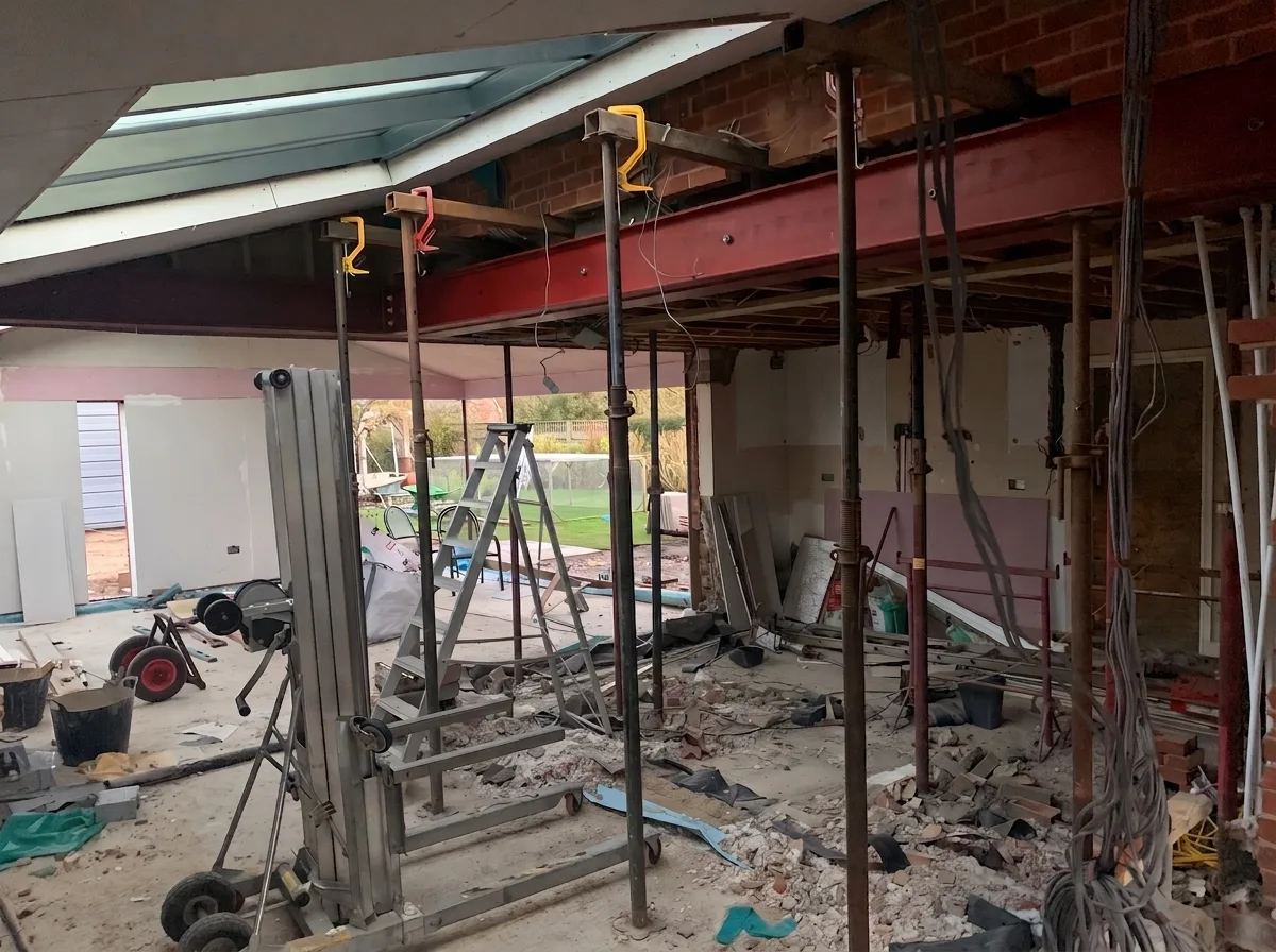 Interior mid-demolition with numerous adjustable steel props supporting a painted I-beam, debris across the concrete floor, a rooflight to one side and garden visible through the open wall.