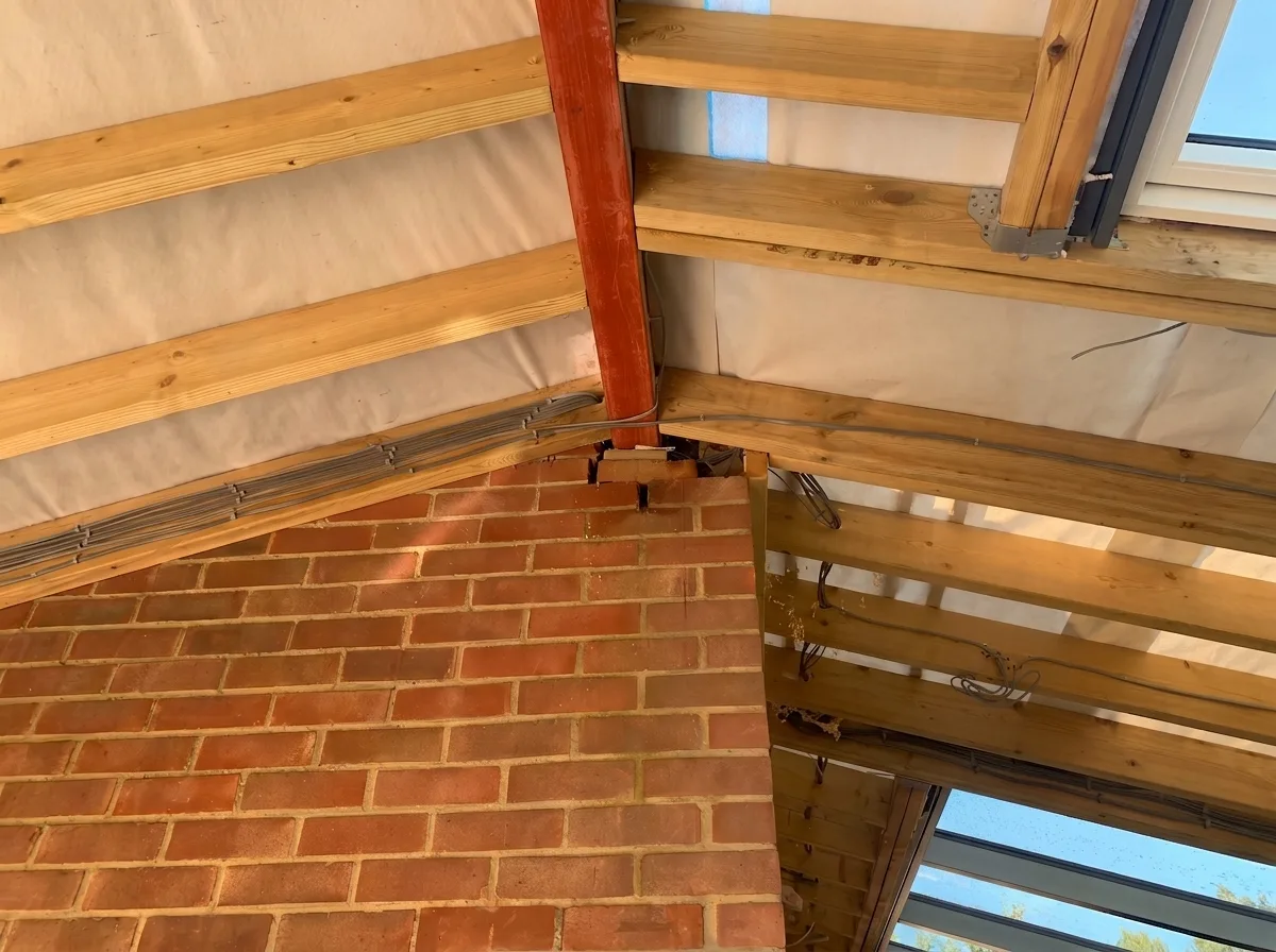 Interior view where a red brick wall meets a timber roof structure, a painted steel member bedded at the wall head, rafters above a white membrane and grey cables run along the timbers.