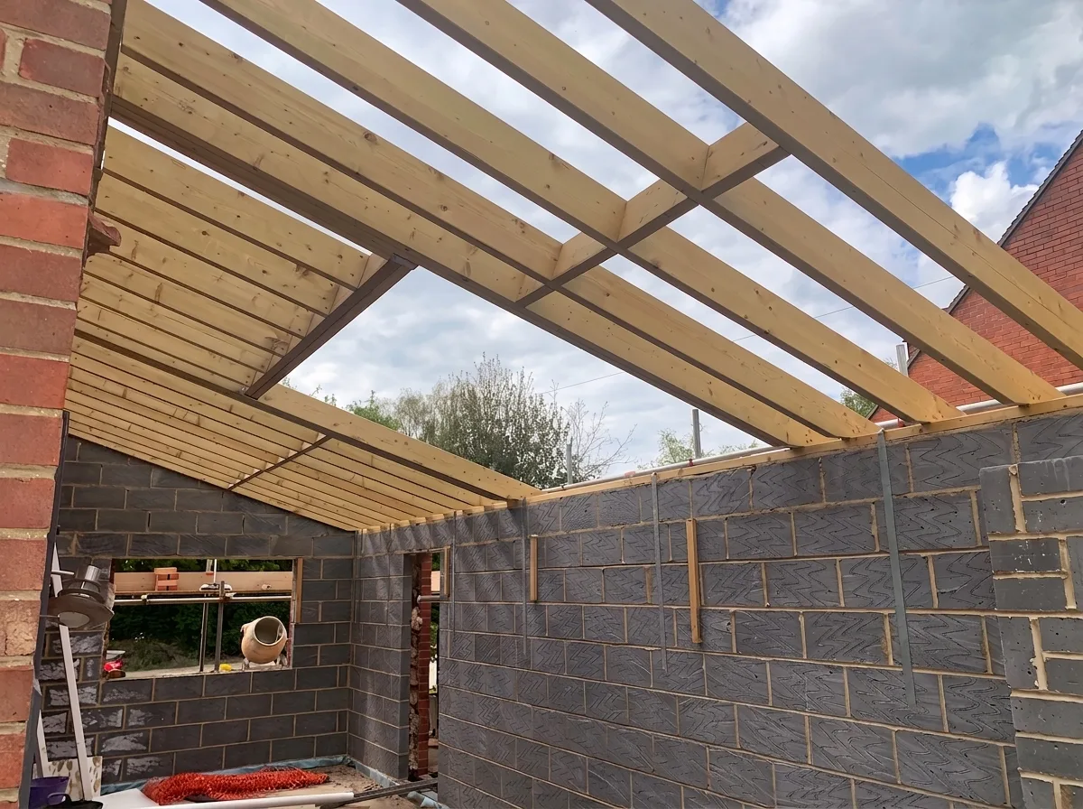 Interior view of an unfinished extension with grey blockwork walls and a new timber roof frame, doubled timbers framing a single window aperture, open sky visible beyond the rafters.