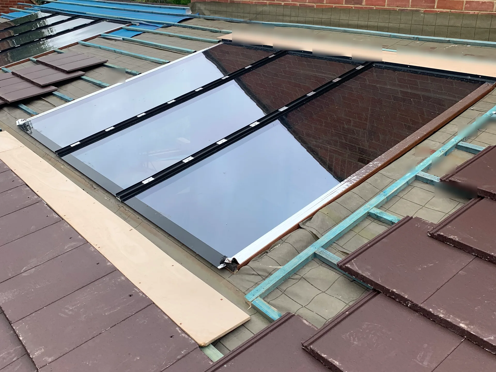 Large multi-panel roof glazing installed into a pitched roof, surrounded by roofing underlay, timber battens and partially laid clay tiles.