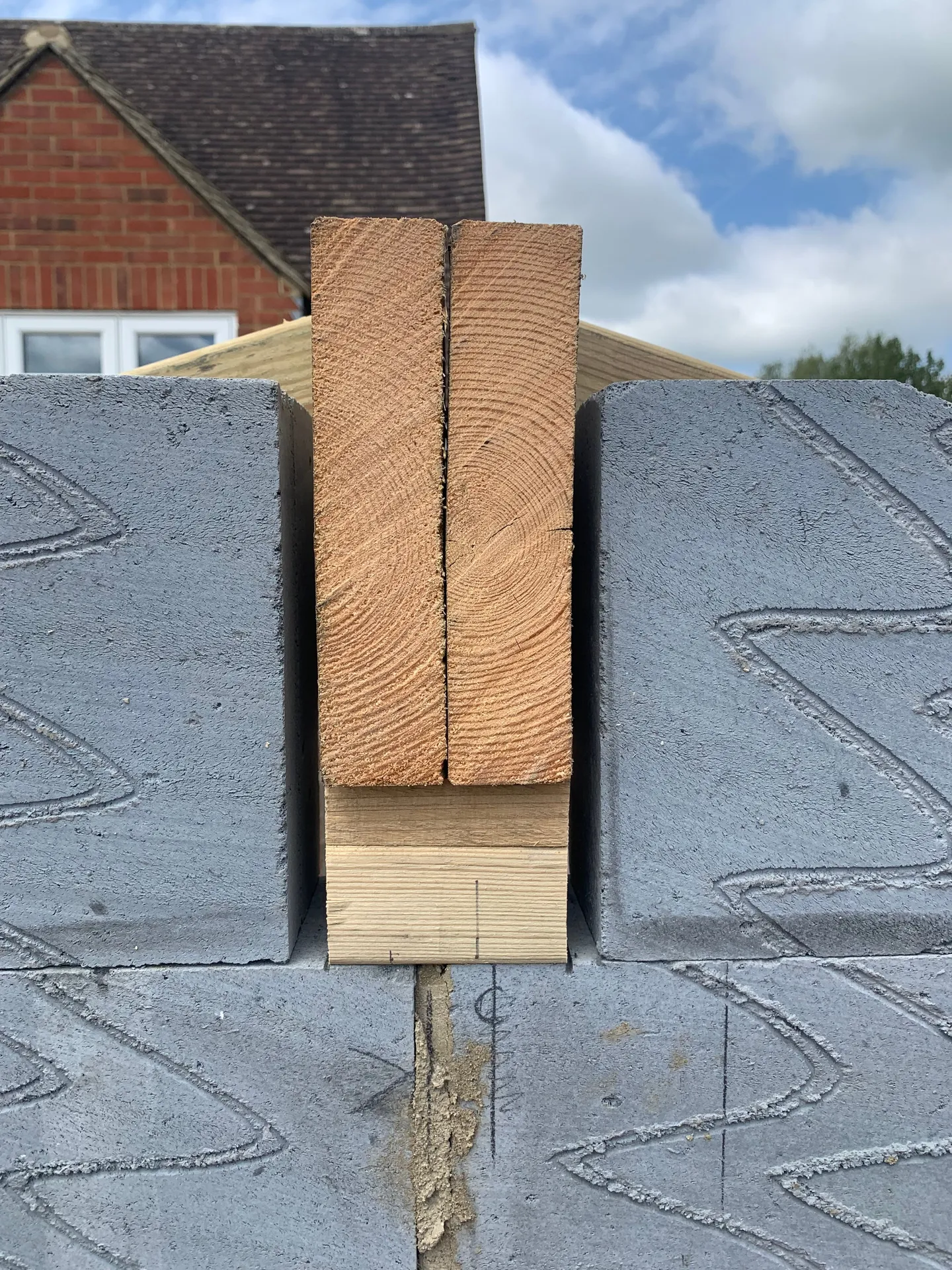Two rough sawn timber planks standing vertically in a recess in a grey blockwork wall, packed at the base with smaller timber offcuts, the roof of a brick house behind them.
